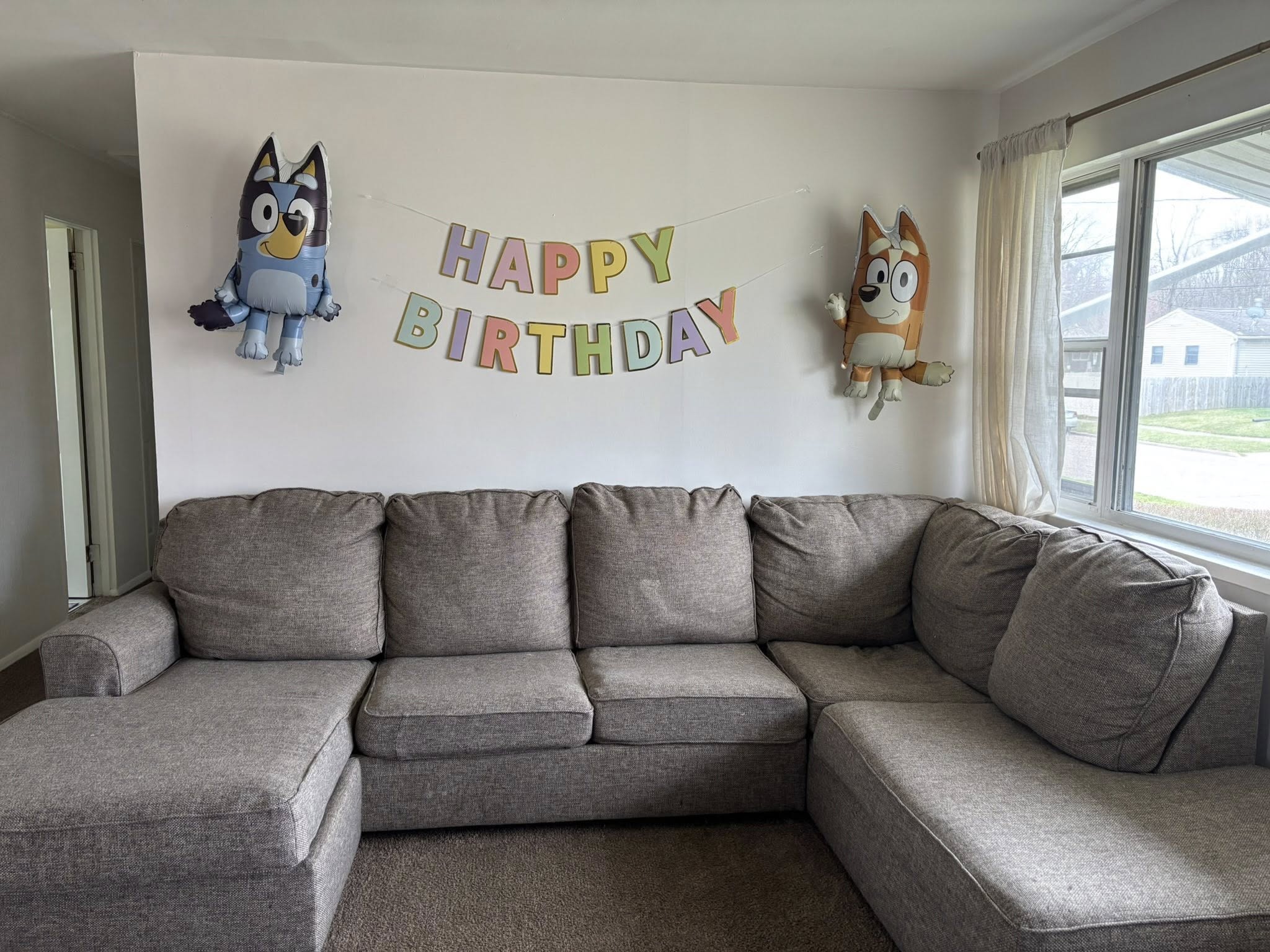 Living room wall decorated with Happy Birthday banner and Bluey and Bingo character balloons above a sectional sofa.