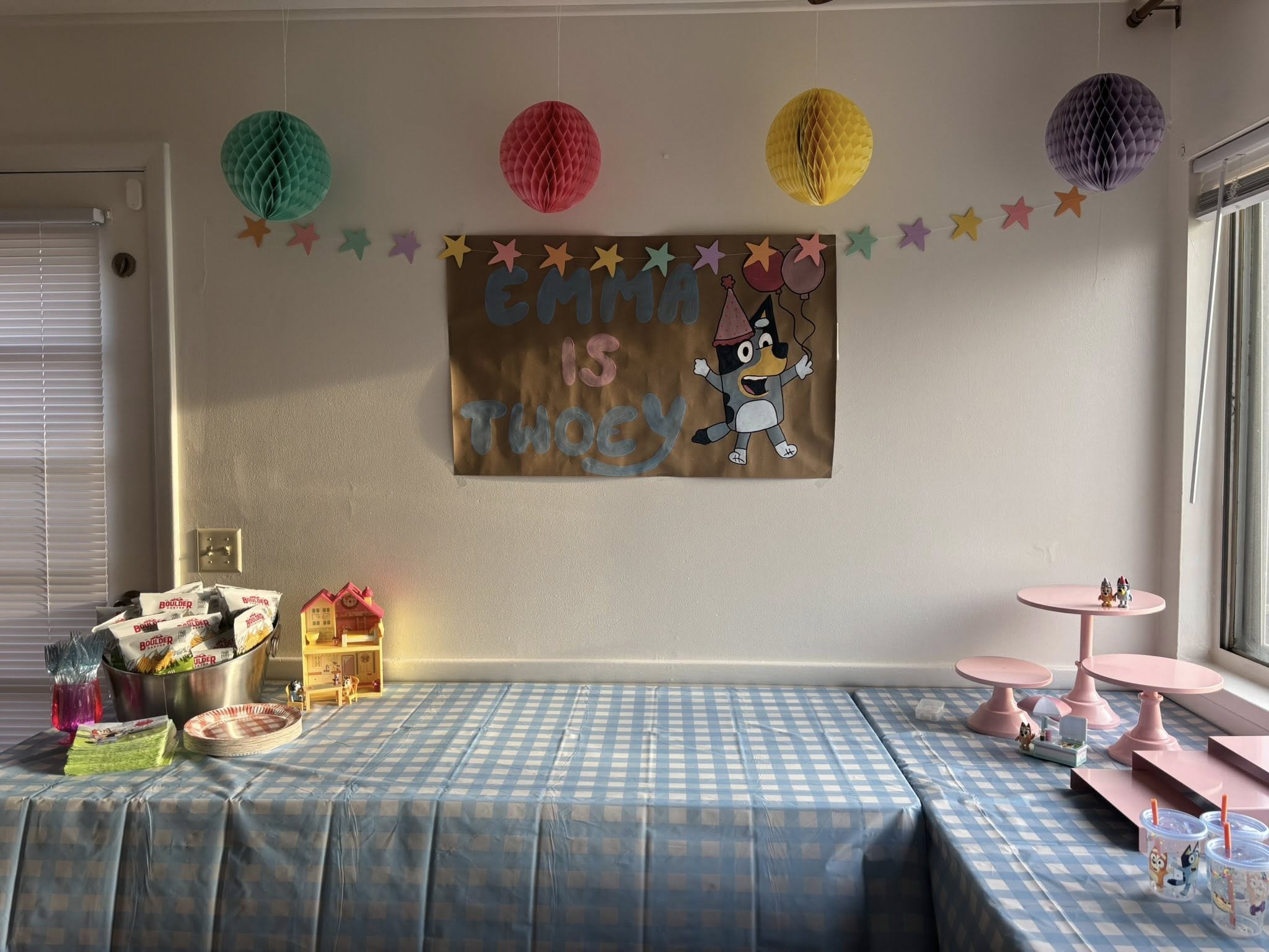 Bluey-themed second birthday party setup with a sign reading Emma Is Twoey, hanging honeycomb decorations, and blue gingham tablecloths.