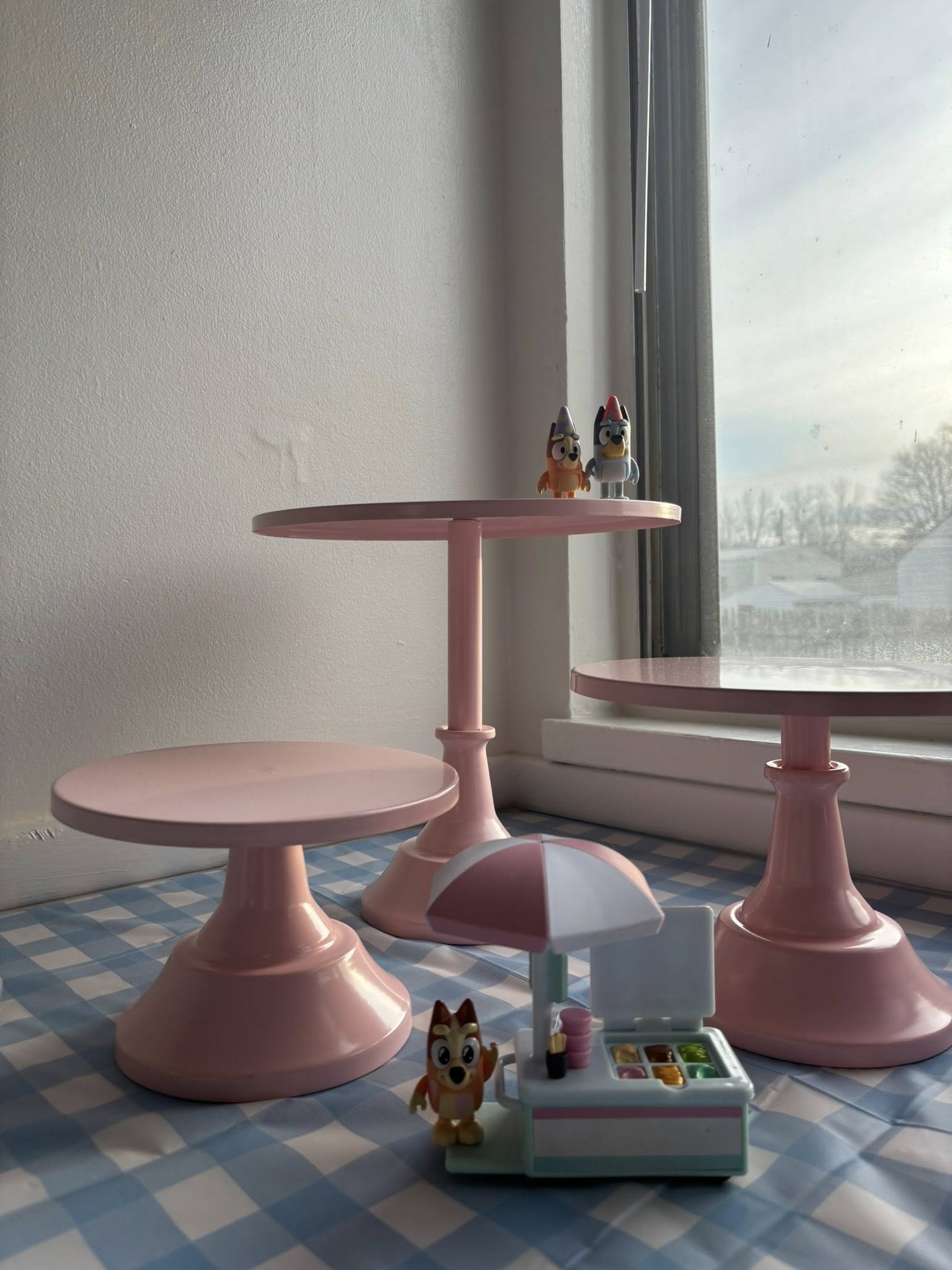 Set of three pink tiered cake stands on a blue and white checkered surface with small toy figurines.