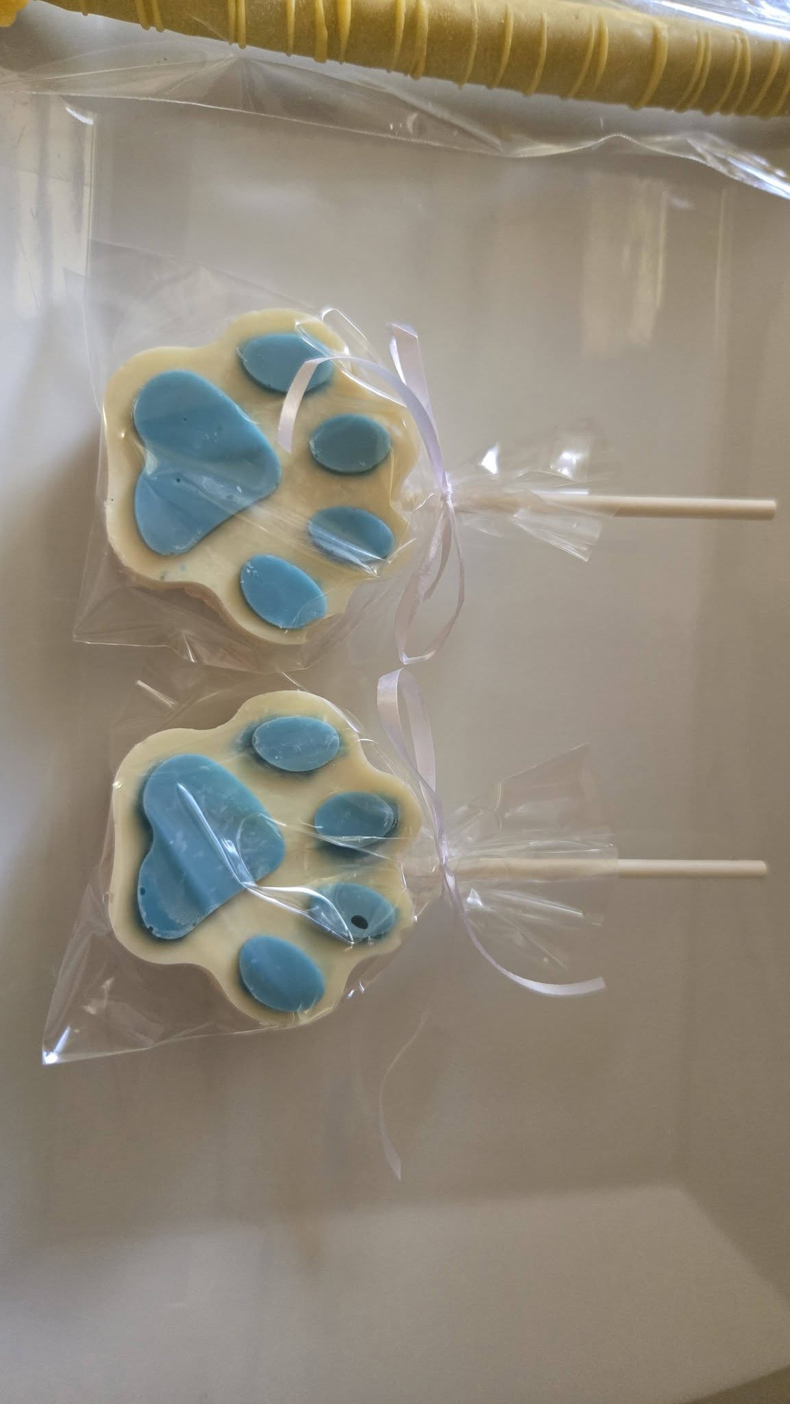 Two white and blue puppy paw print chocolate lollipops wrapped in cellophane with a ribbon.