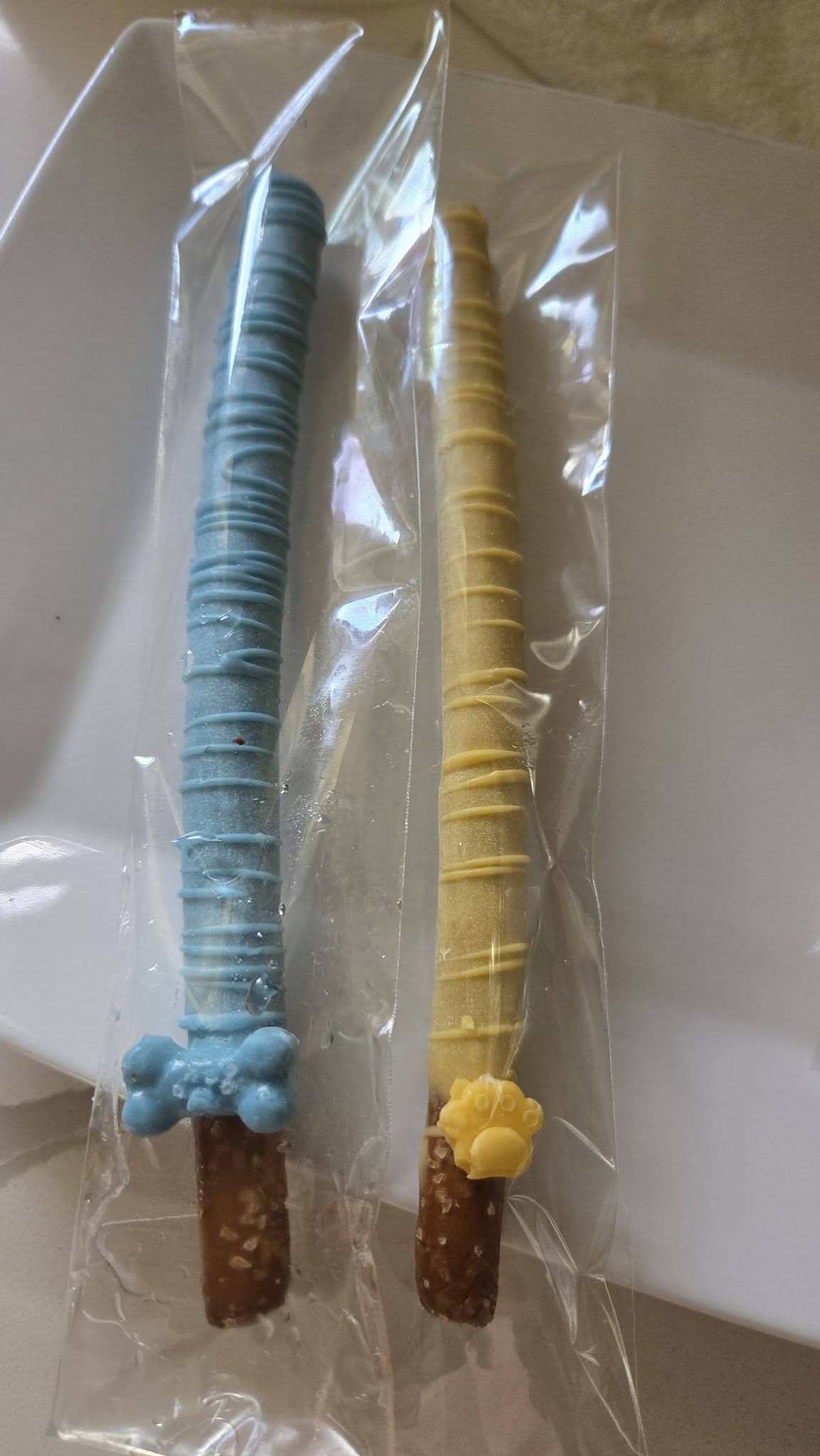 Two individually wrapped pretzel rods dipped in blue and yellow candy, decorated with paw and bone shaped icing.