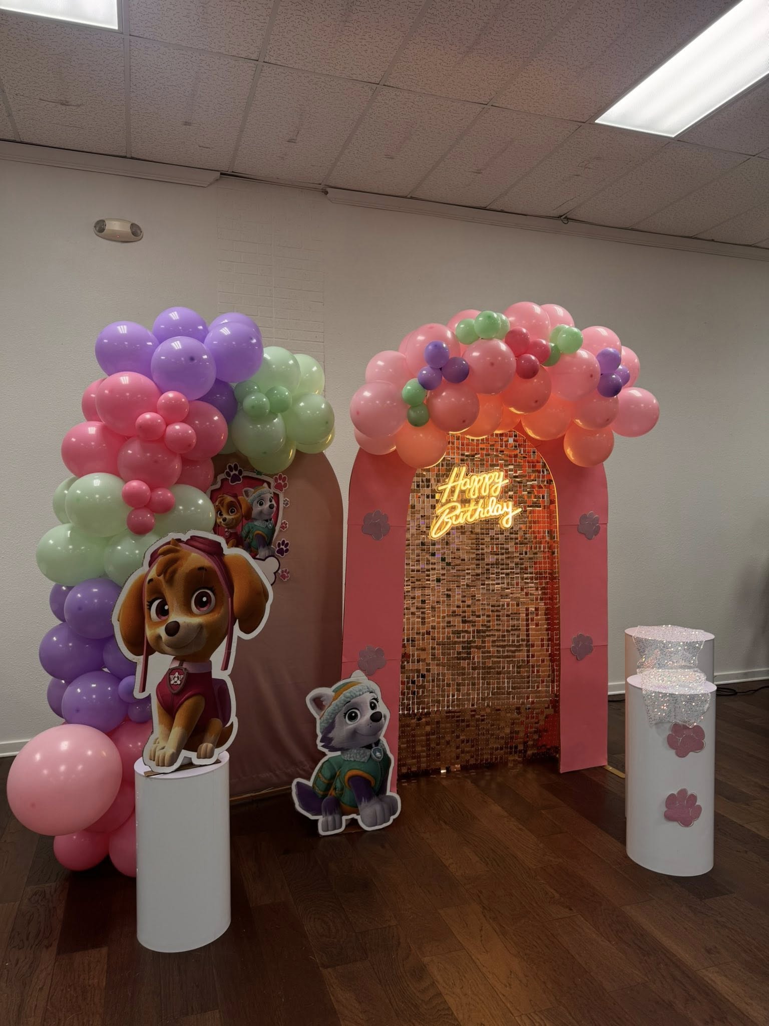 Paw Patrol themed birthday party backdrop with pink arch, gold shimmer wall, Happy Birthday neon sign, and balloons.