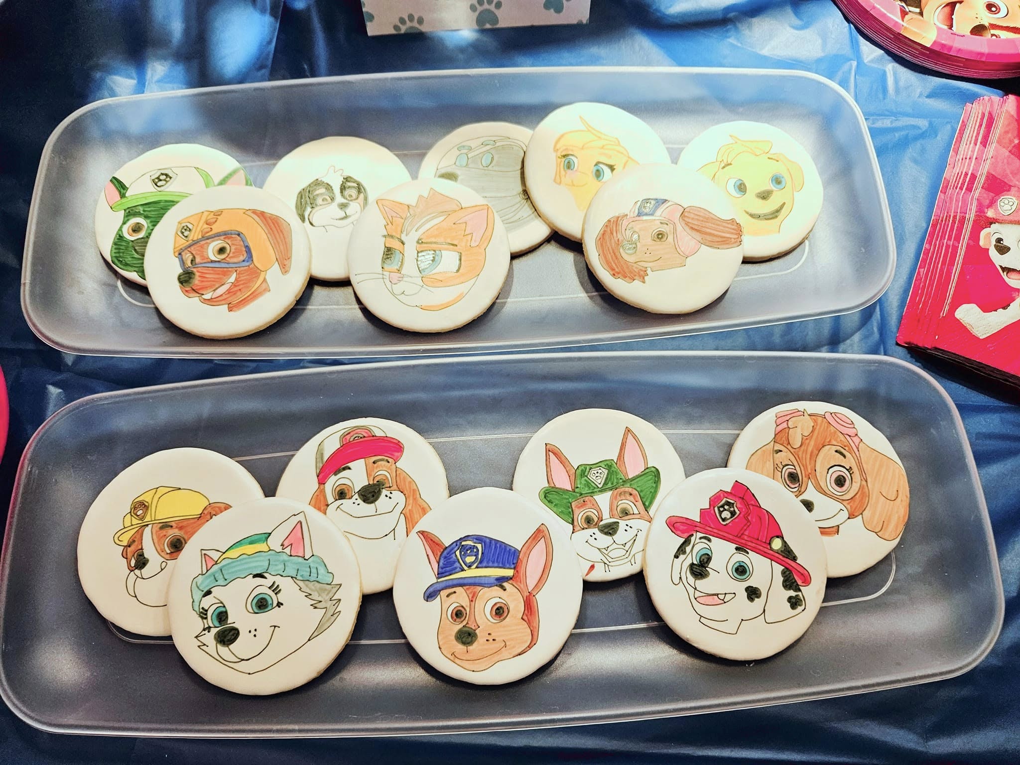 Two clear trays of round Paw Patrol character cookies on a blue party tablecloth.