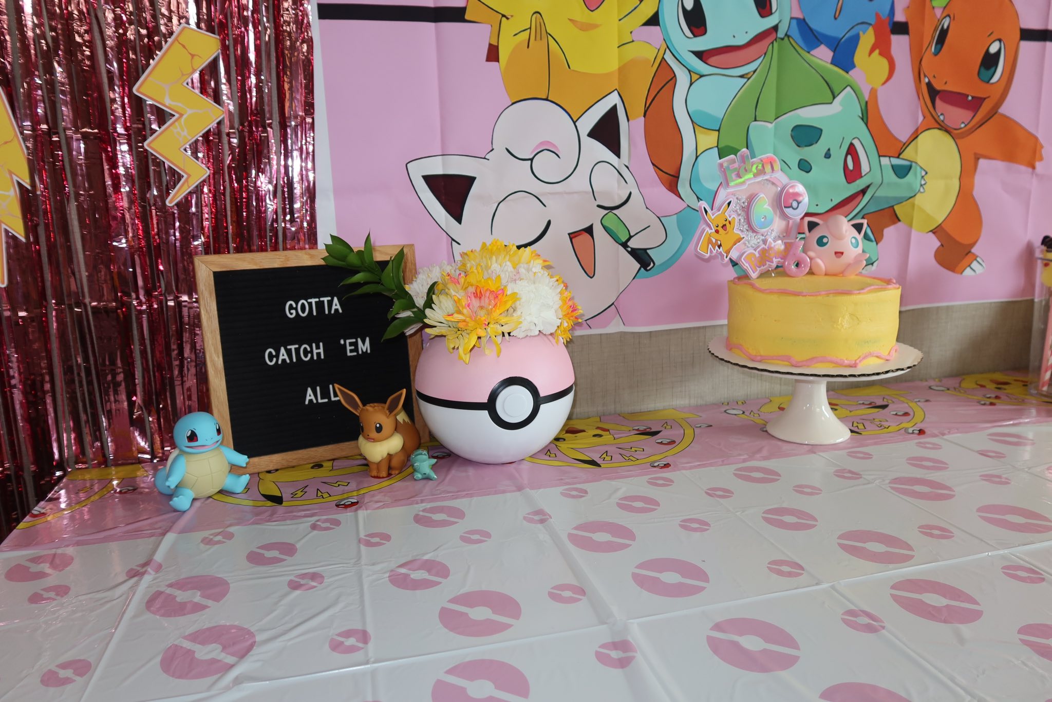 Pokemon themed birthday party table with a yellow cake, Poké Ball flower vase, and letter board sign.