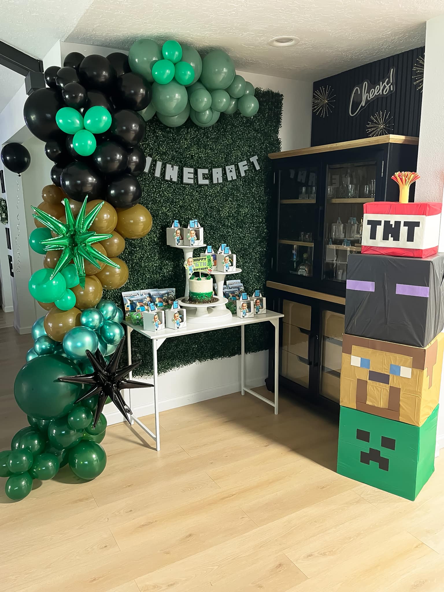 Minecraft themed birthday party setup with a balloon garland backdrop, cake, and stacked block party decorations.