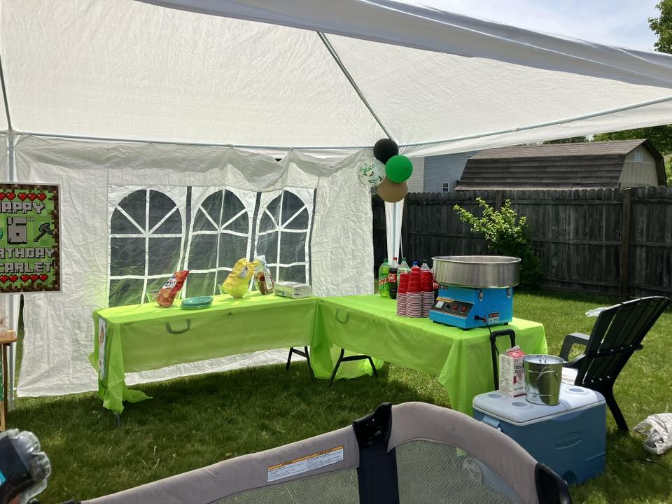 Outdoor birthday party setup with a green tablecloth, snacks, and a cotton candy machine under a white tent.