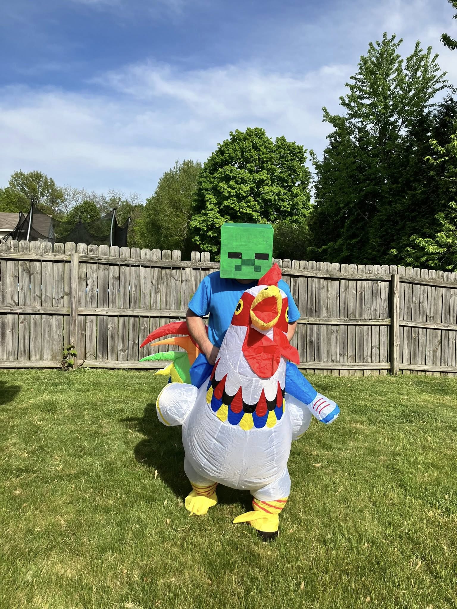 Person wearing an inflatable chicken costume and a square green Creeper mask in a backyard.