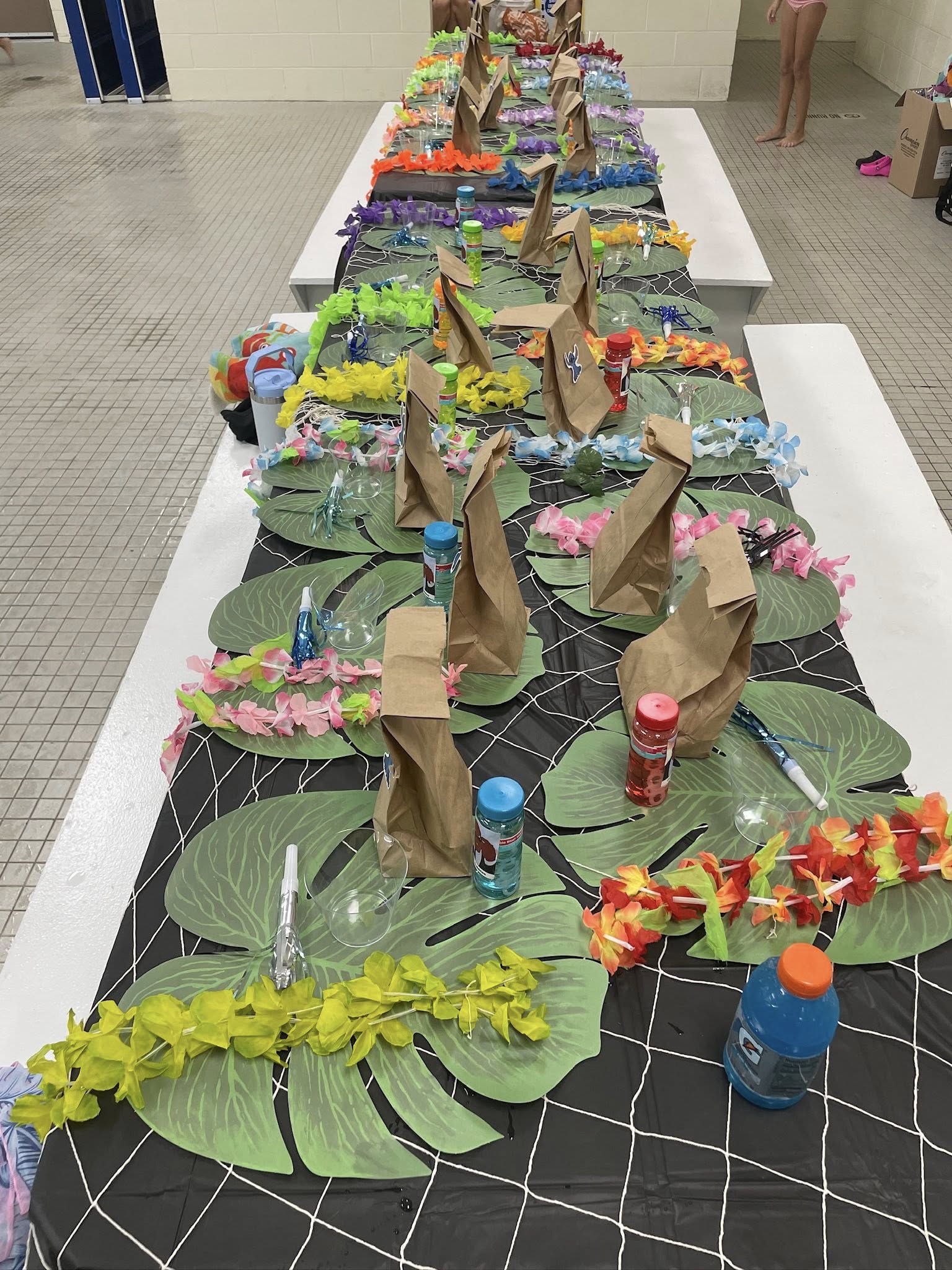 Long table set for a tropical themed party with brown paper bags, colorful leis, and leaf-shaped placemats.