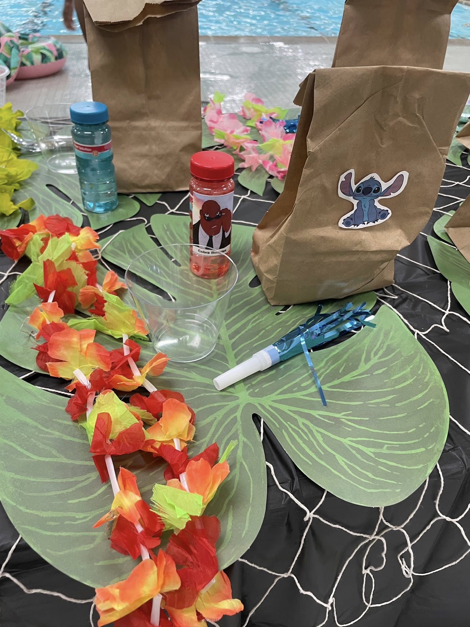 Luau-themed party favors including Stitch stickers on paper bags, colorful flower leis, and bubbles on a tropical table setting.