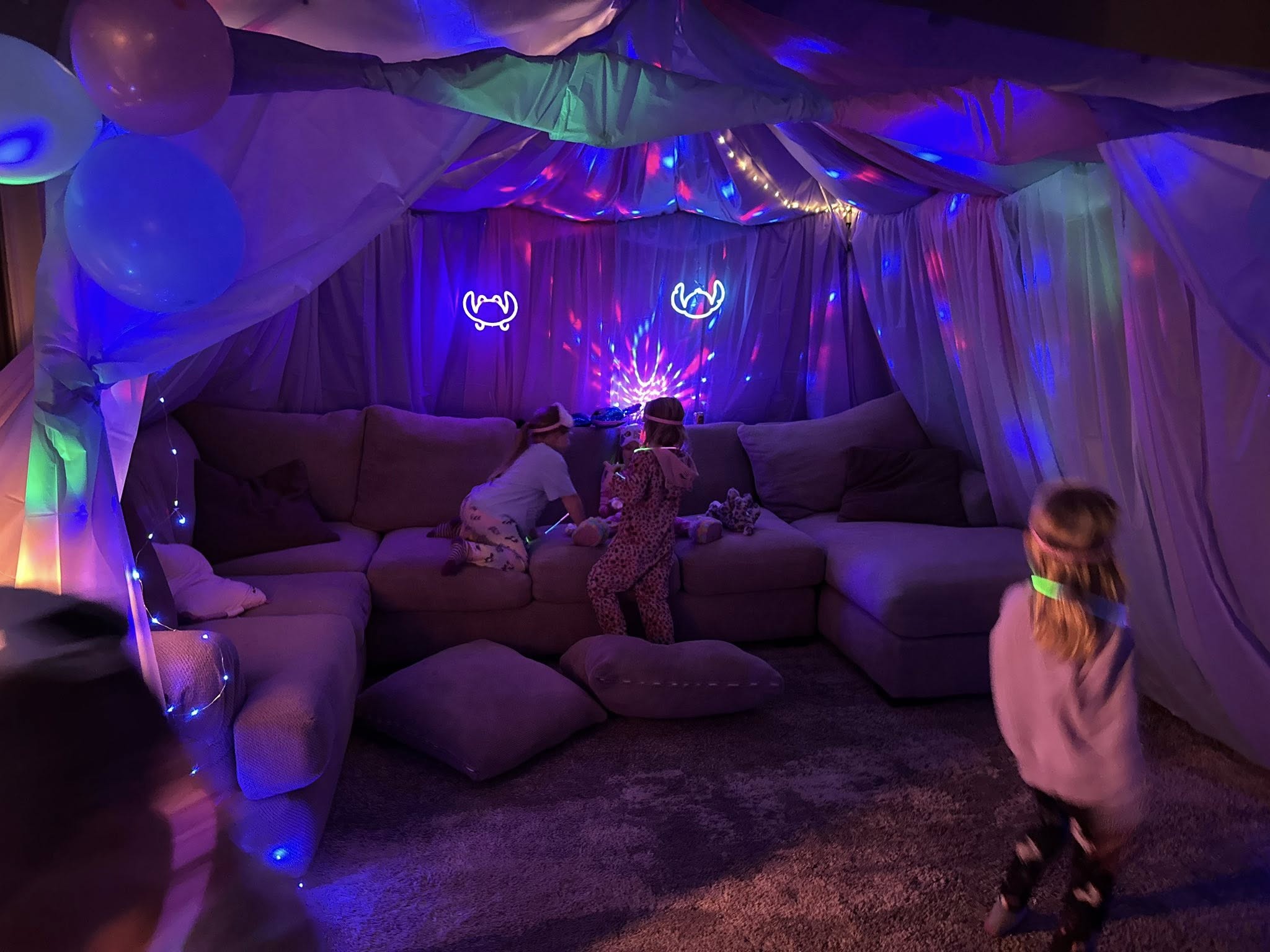 Children playing in a glowing fort made of blankets and fairy lights set up over a living room sectional sofa.