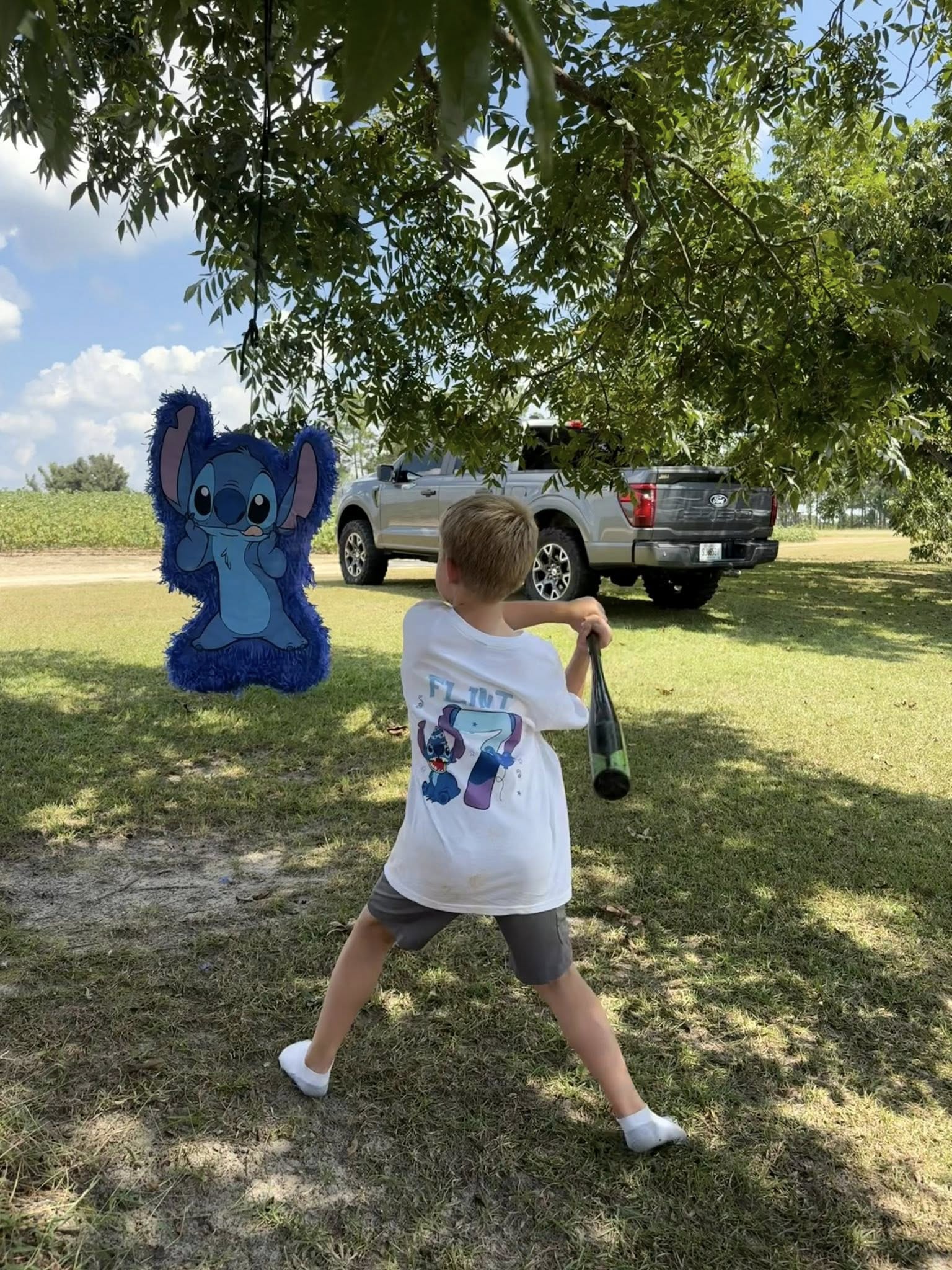 Young boy wearing a personalized Stitch birthday shirt swinging at a Stitch pinata under a tree.