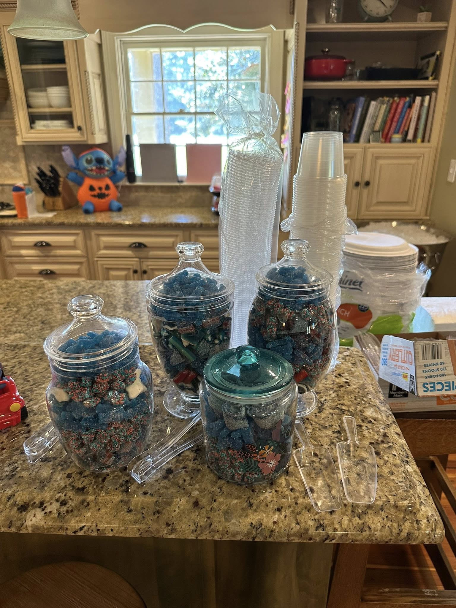 Glass candy jars filled with blue gummy treats on a kitchen counter with sets of clear plastic cups and cutlery