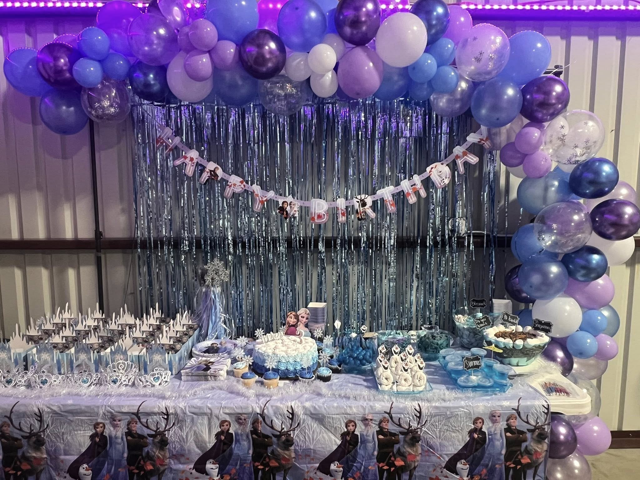Frozen-themed dessert table with blue and purple balloon arch and Happy Birthday banner.
