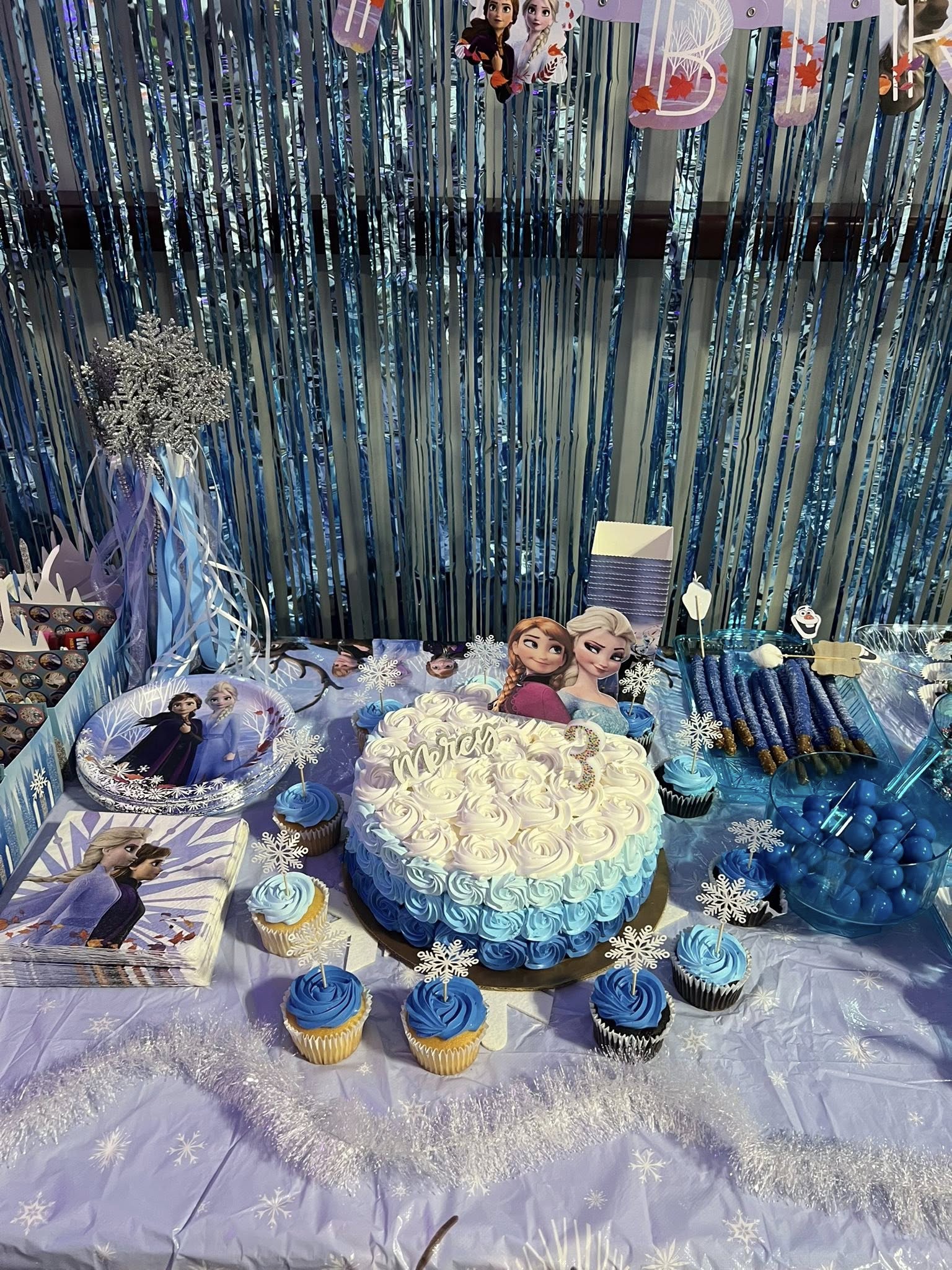 Frozen themed birthday party table with blue ombre cake, matching cupcakes, plates, and napkins against a blue fringe backdrop.