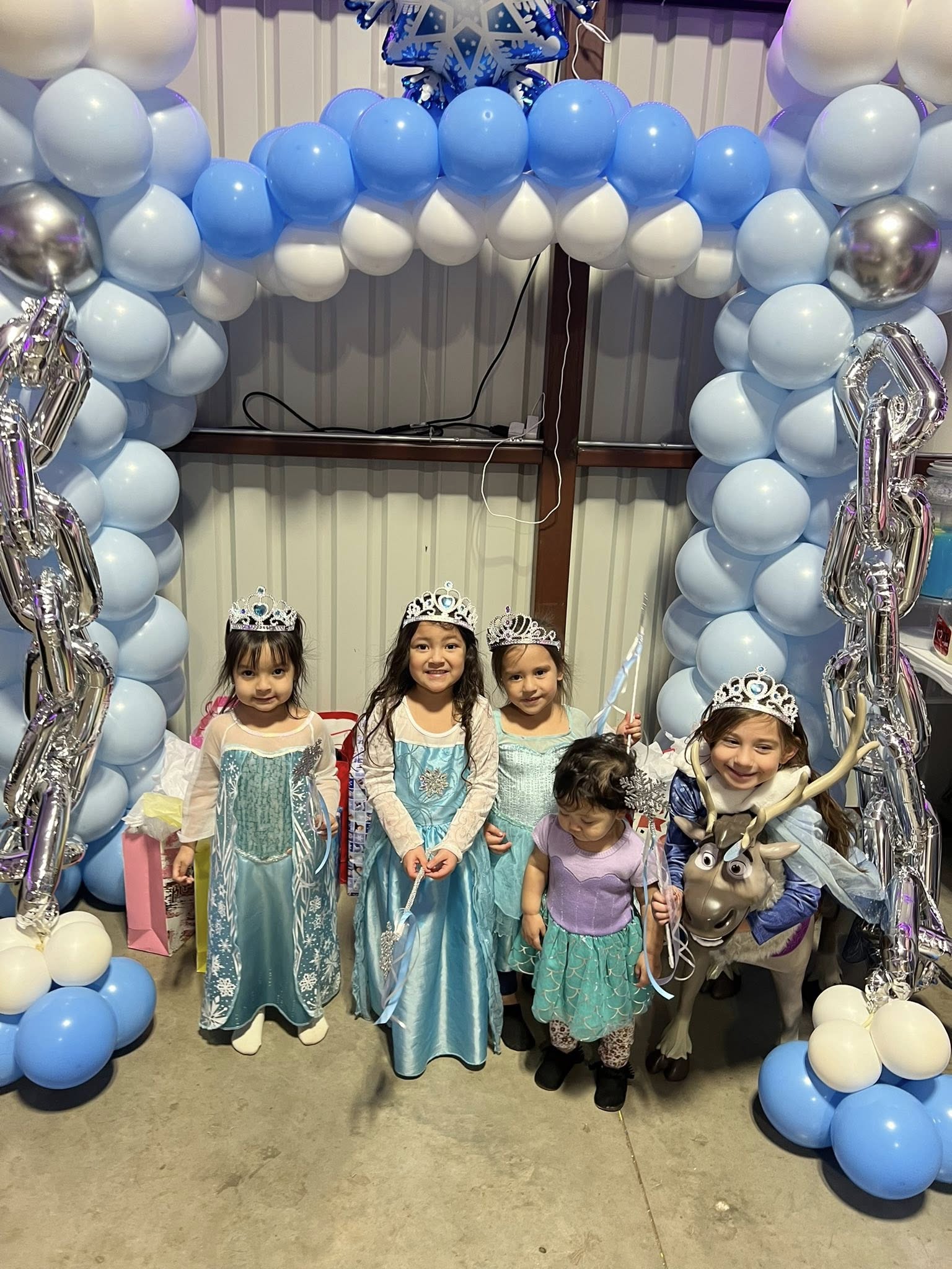 Four young girls in princess costumes posing under a blue and white balloon arch for a birthday party.