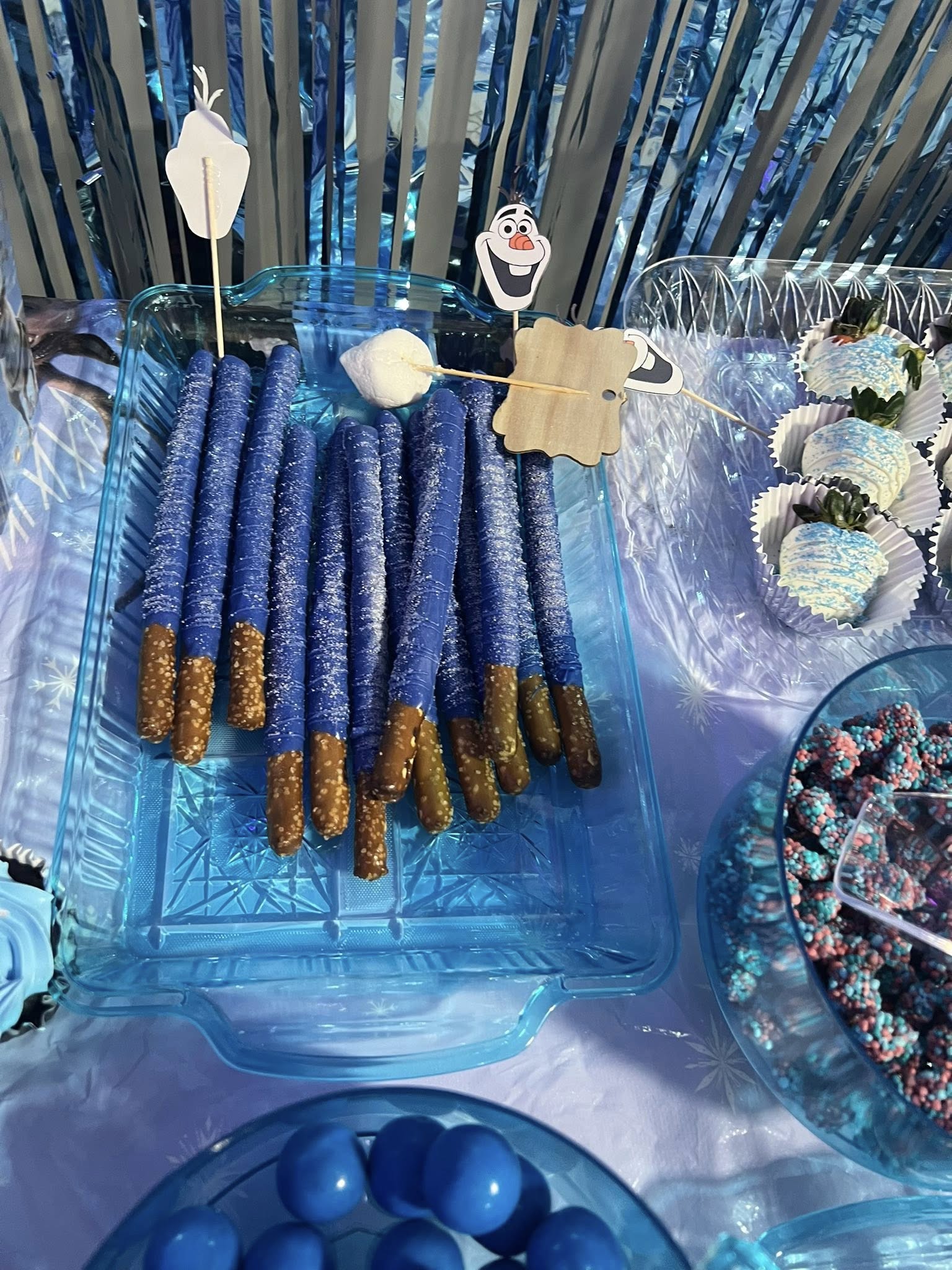 Table with blue chocolate-dipped pretzel rods, white chocolate-covered strawberries, and blue candies for a Frozen theme party.