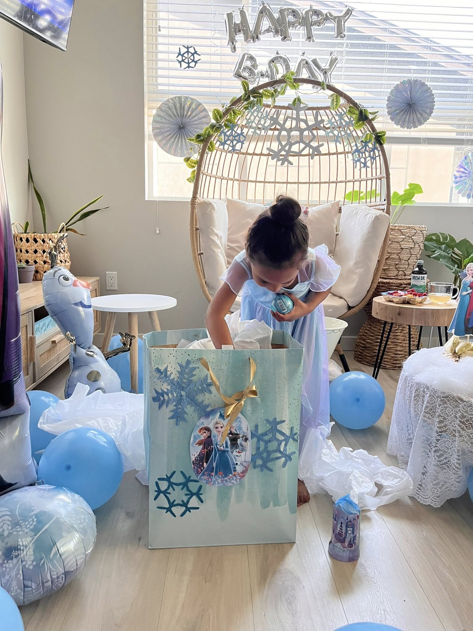 Young girl in a princess dress opening a large Frozen-themed gift bag surrounded by birthday decorations