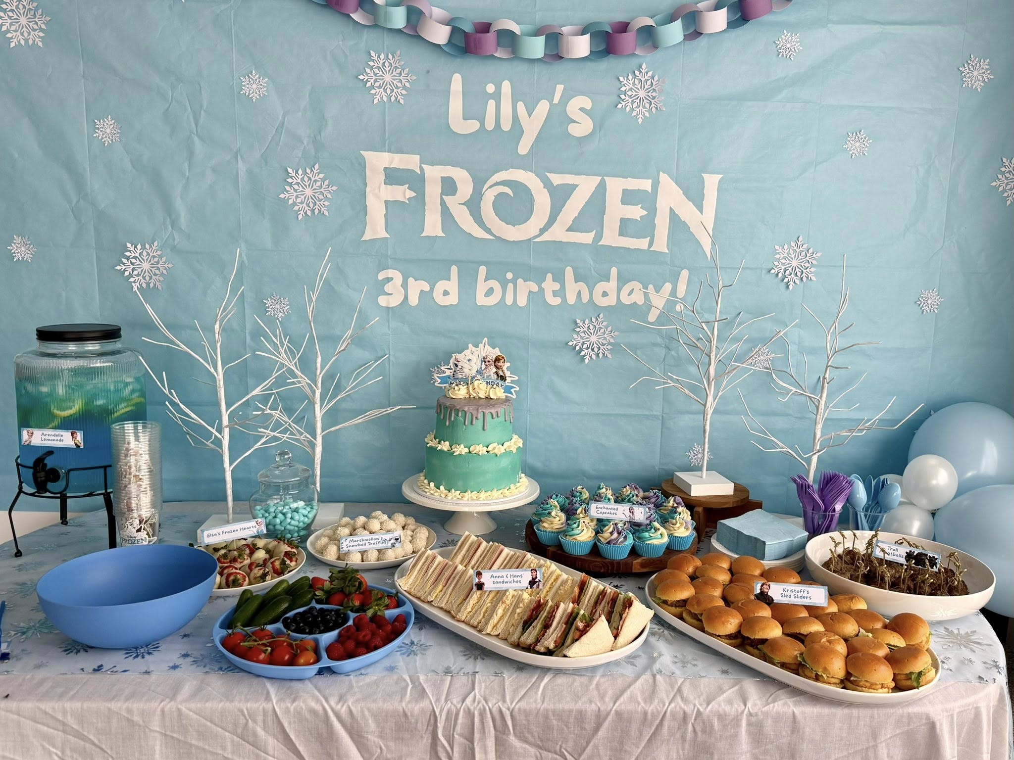 Frozen-themed dessert and snack table for a third birthday party with cake, cupcakes, and sandwiches.