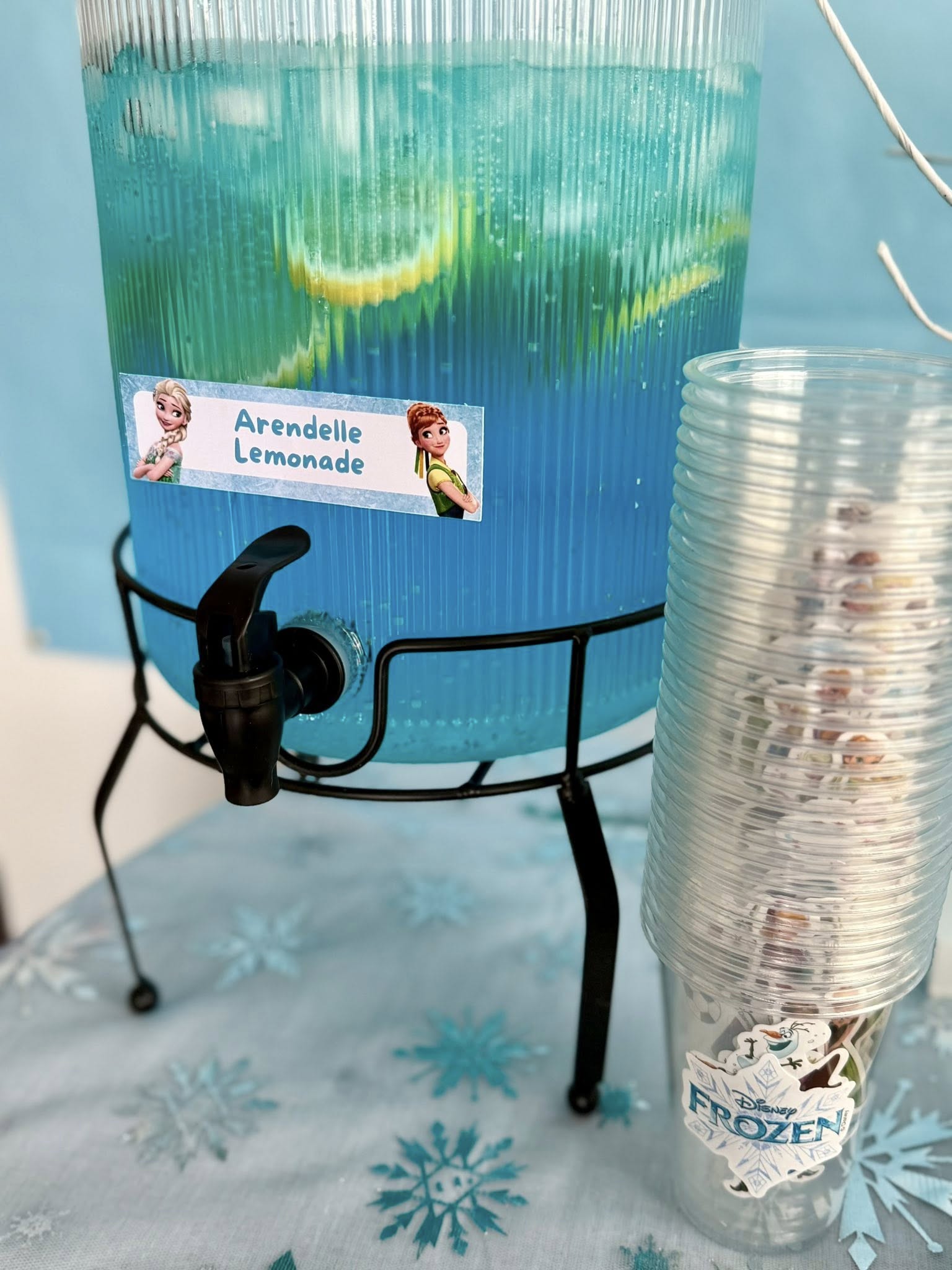 Beverage dispenser filled with blue Arendelle Lemonade and a stack of Frozen-themed plastic cups.