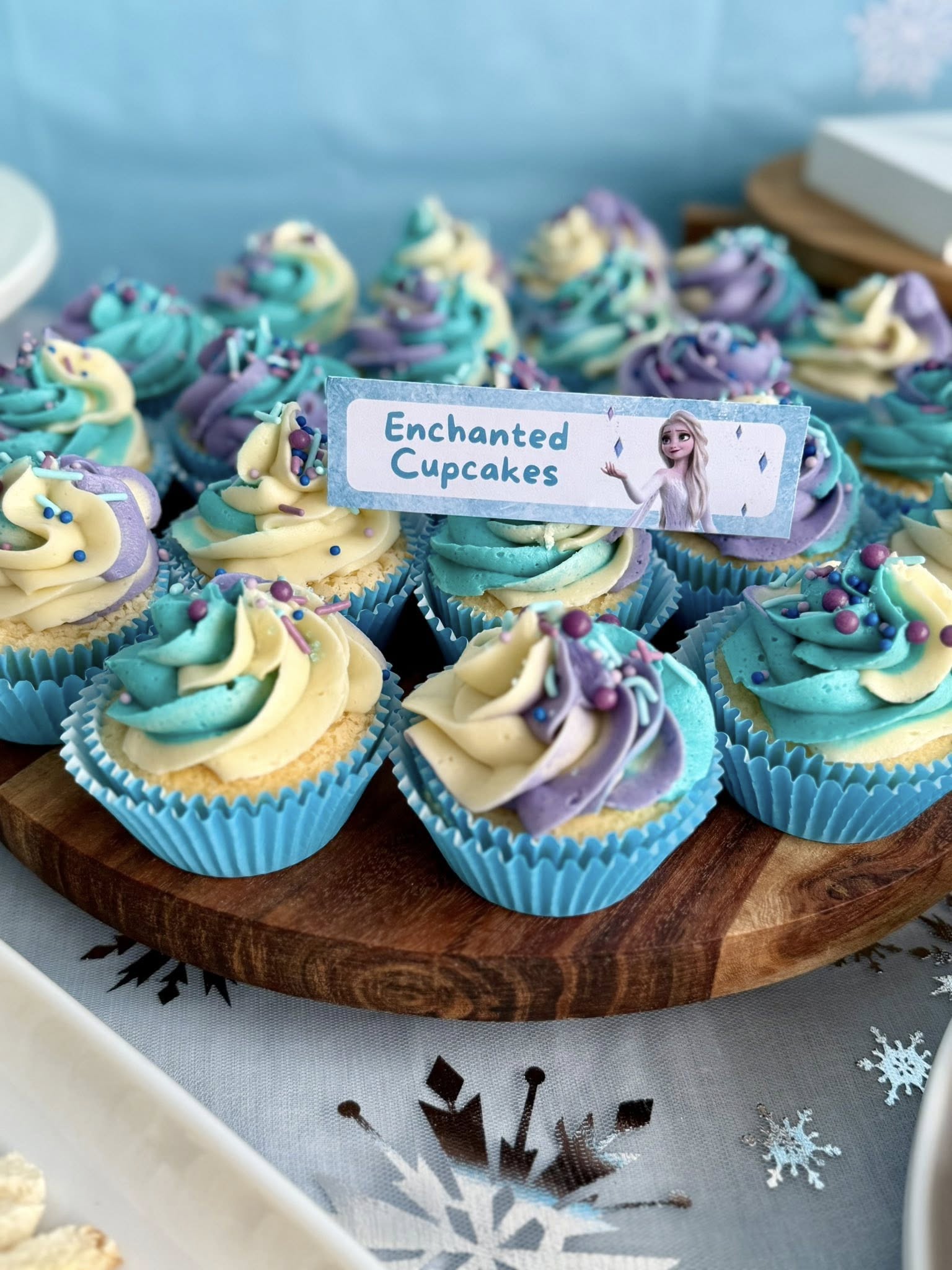 Tray of cupcakes with blue, purple, and white swirled frosting, topped with sprinkles and an Enchanted sign.