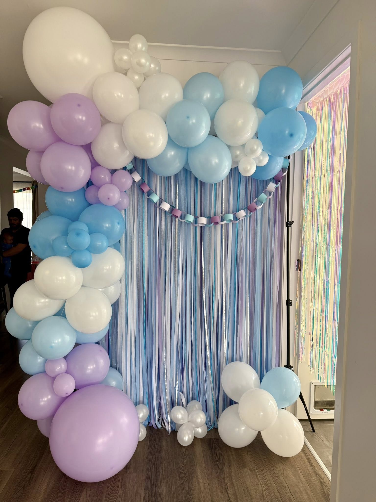 Party balloon arch and streamer backdrop in shades of pastel blue, white, and purple.