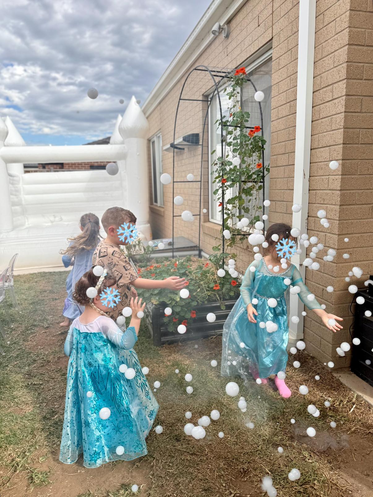 Children playing with floating bubbles in front of a white bouncy castle in a backyard.