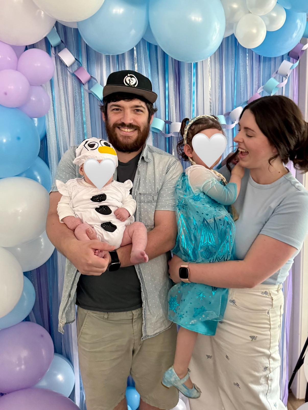Family posing in front of Frozen themed party decorations with the baby dressed as Olaf and child as Elsa.