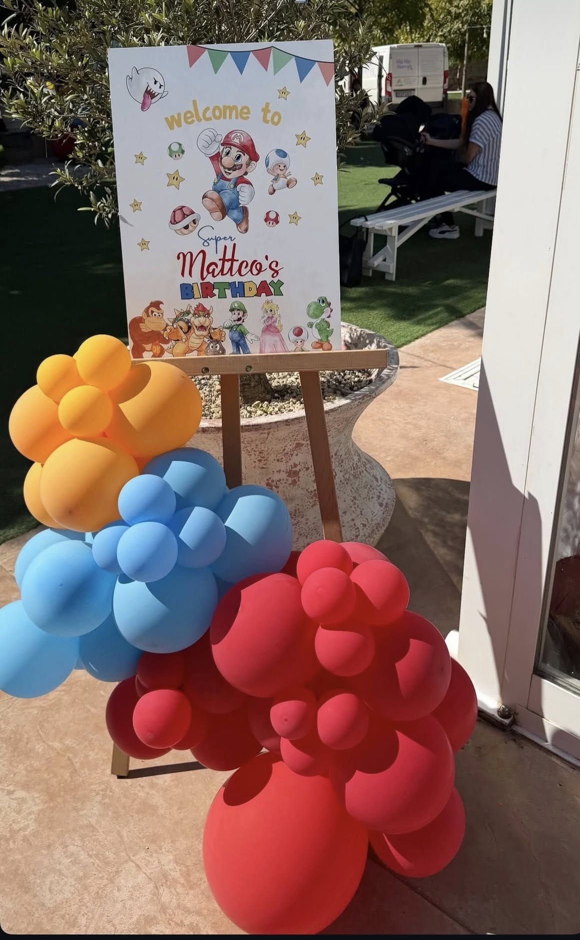 Super Mario themed welcome sign for Matteo's birthday decorated with colorful balloons on a wooden easel