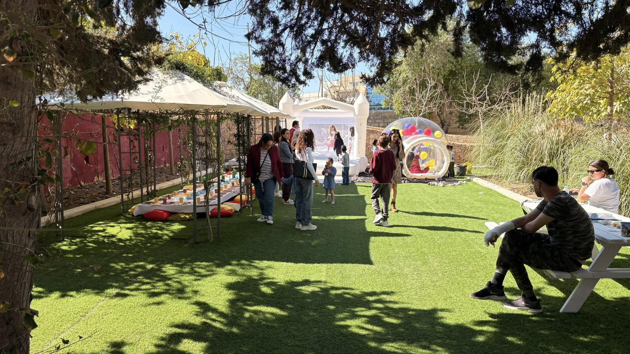 Children and adults at a sunny outdoor party with a white bounce house and a bubble tent filled with balloons.