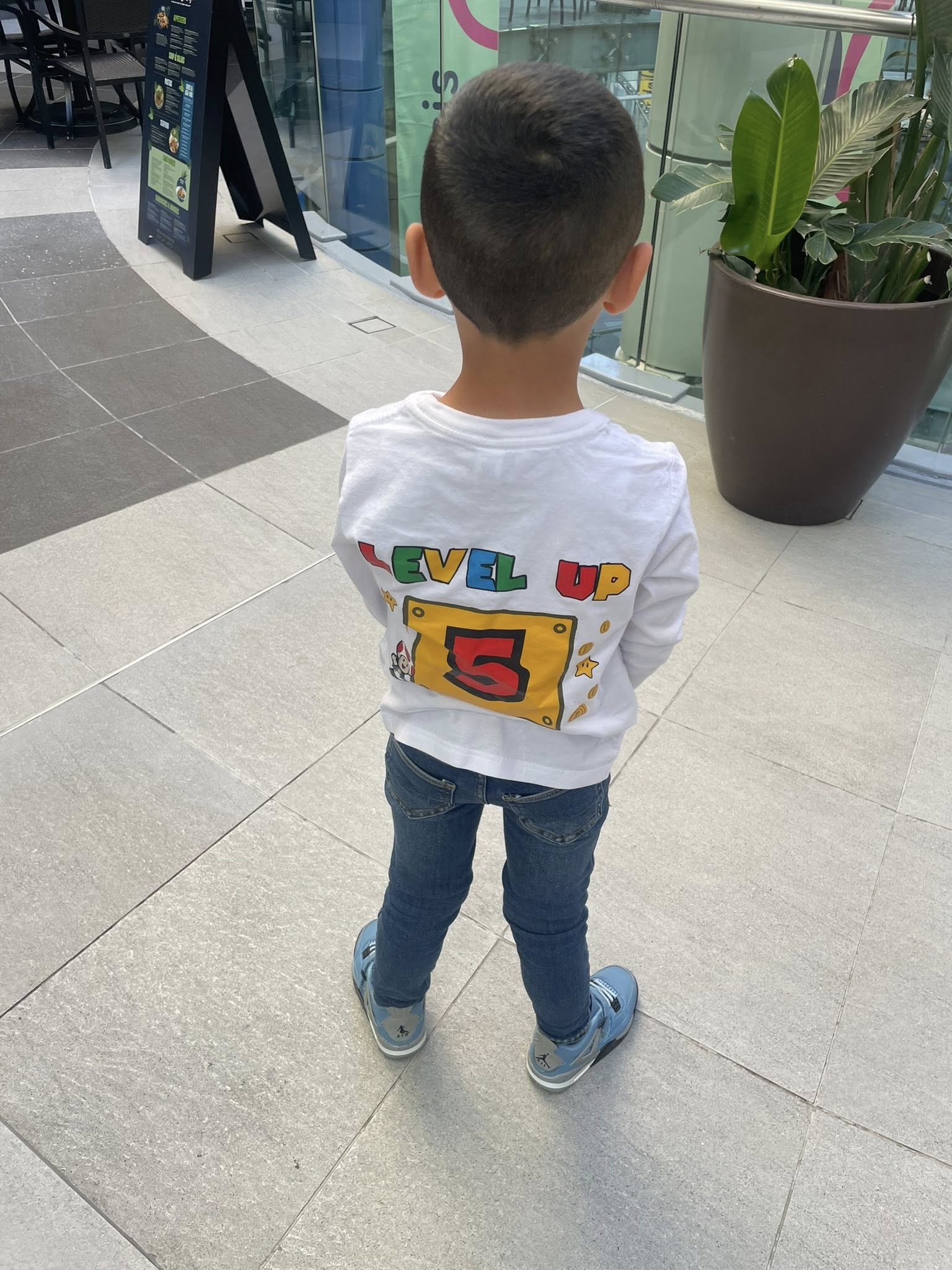 Young boy wearing a white long-sleeve shirt with a Level Up 5 video game themed graphic on the back.