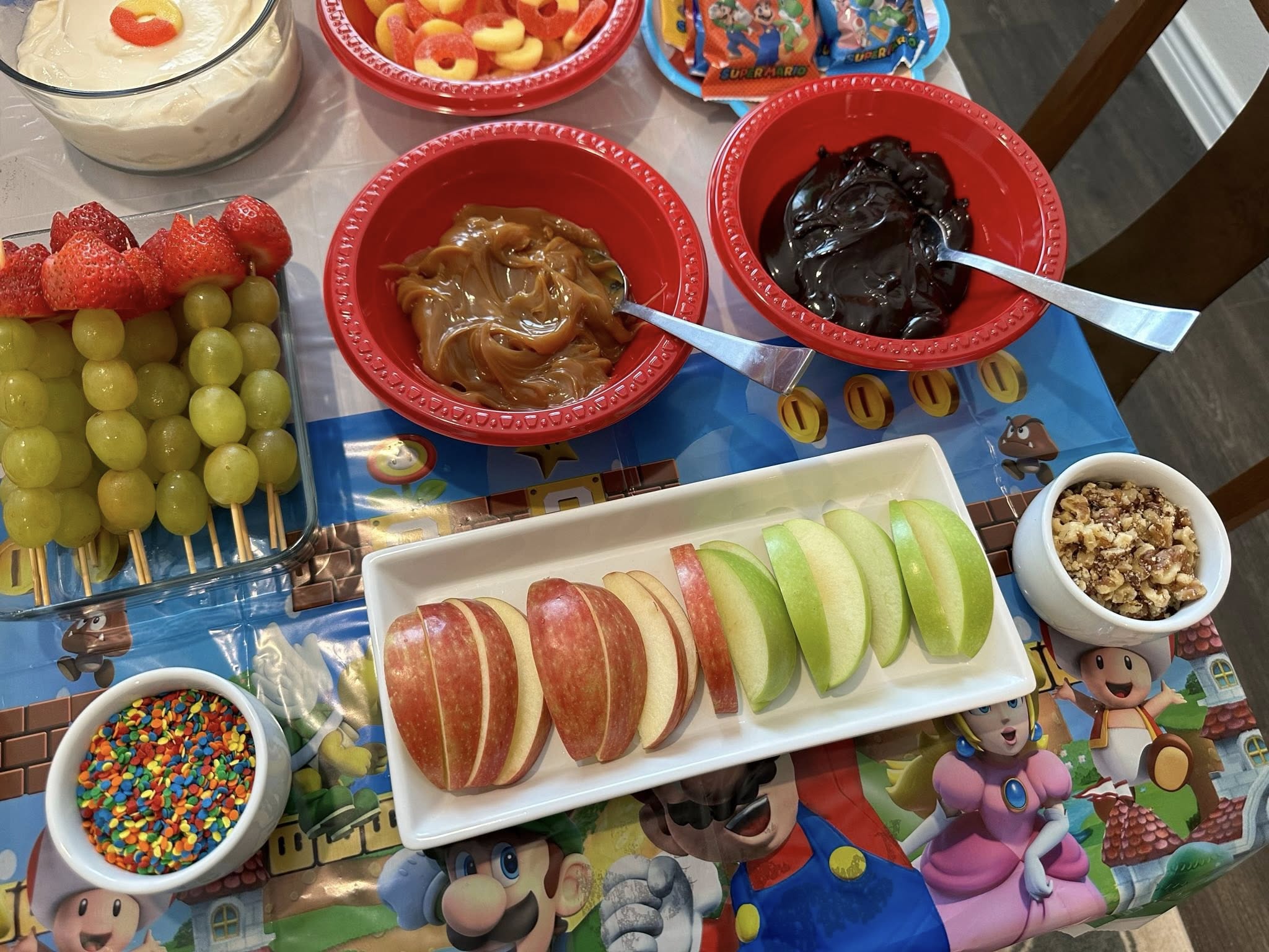 Fruit platter with sliced apples, strawberry-grape skewers, caramel, chocolate dip, sprinkles, and nuts on a Mario tablecloth