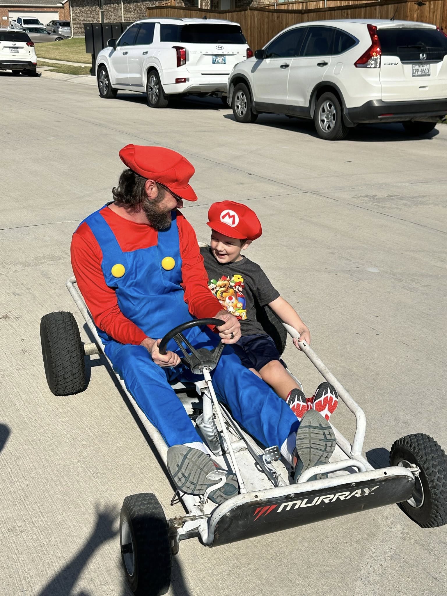 Father and young son dressed as Mario riding a vintage Murray pedal kart together on a suburban street