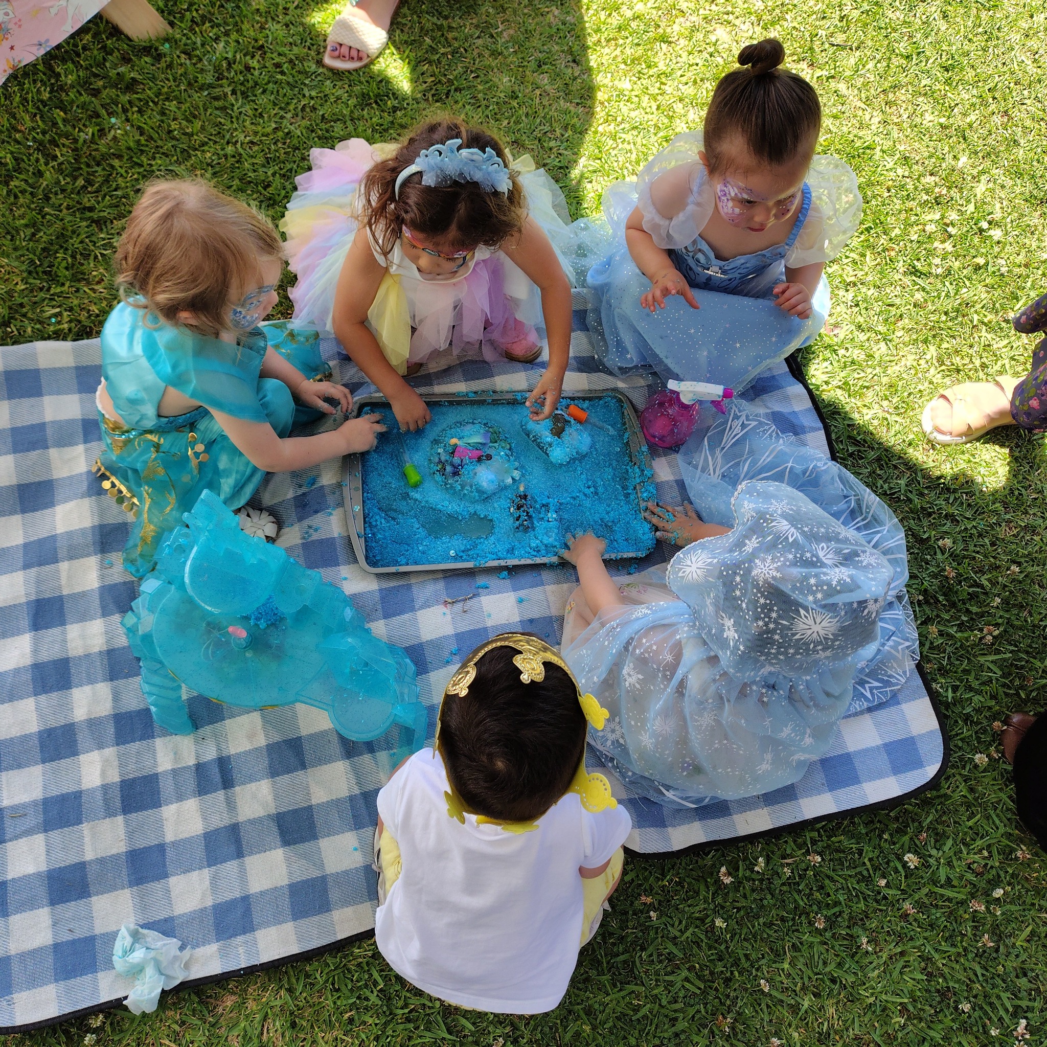 Children in princess costumes playing with blue sensory sand on a picnic blanket outdoors.