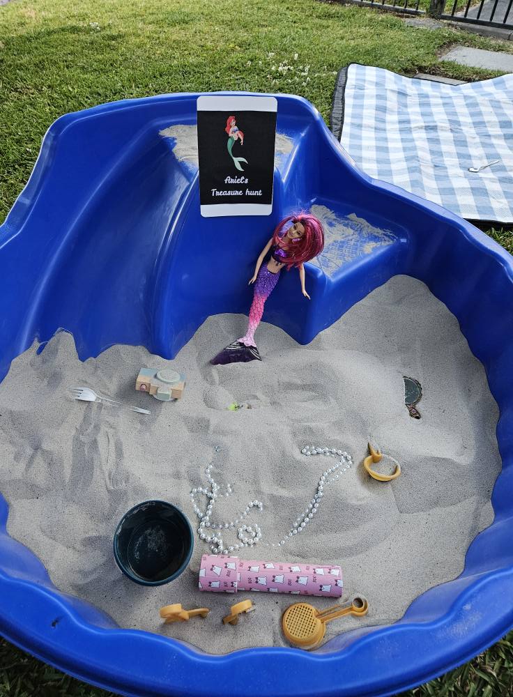 Blue plastic sandpit holding sand, a mermaid doll, a themed treasure hunt sign, jewelry, and various toys.