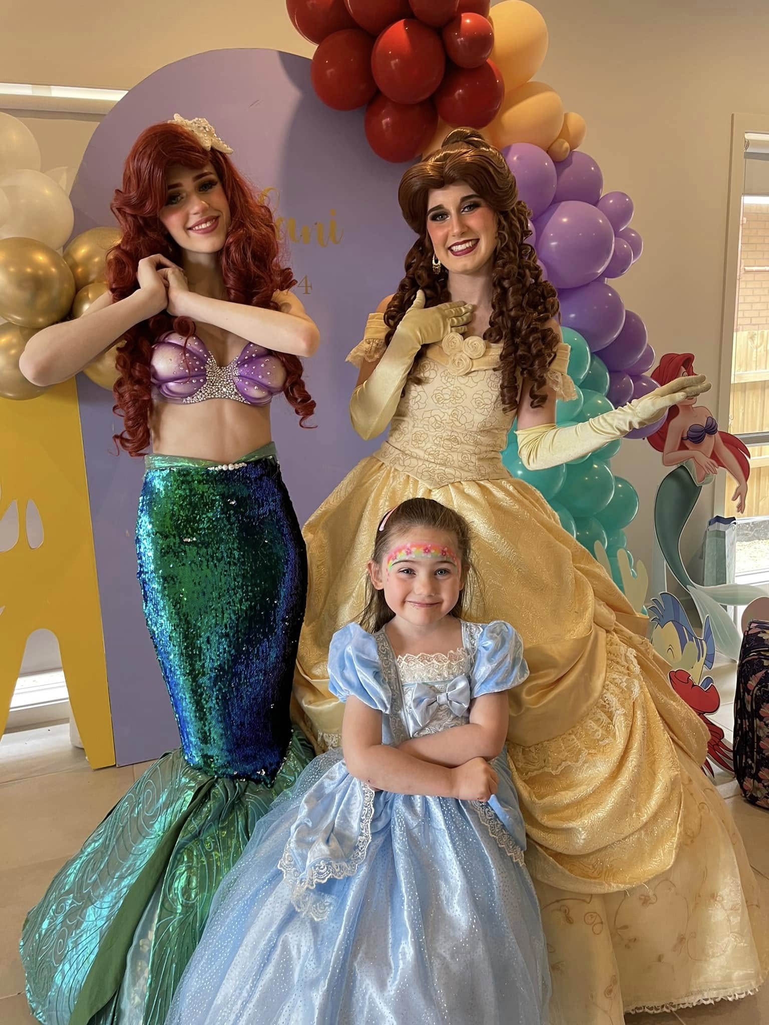 A young girl in a blue princess dress posing with performers dressed as Ariel and Belle at a birthday party.