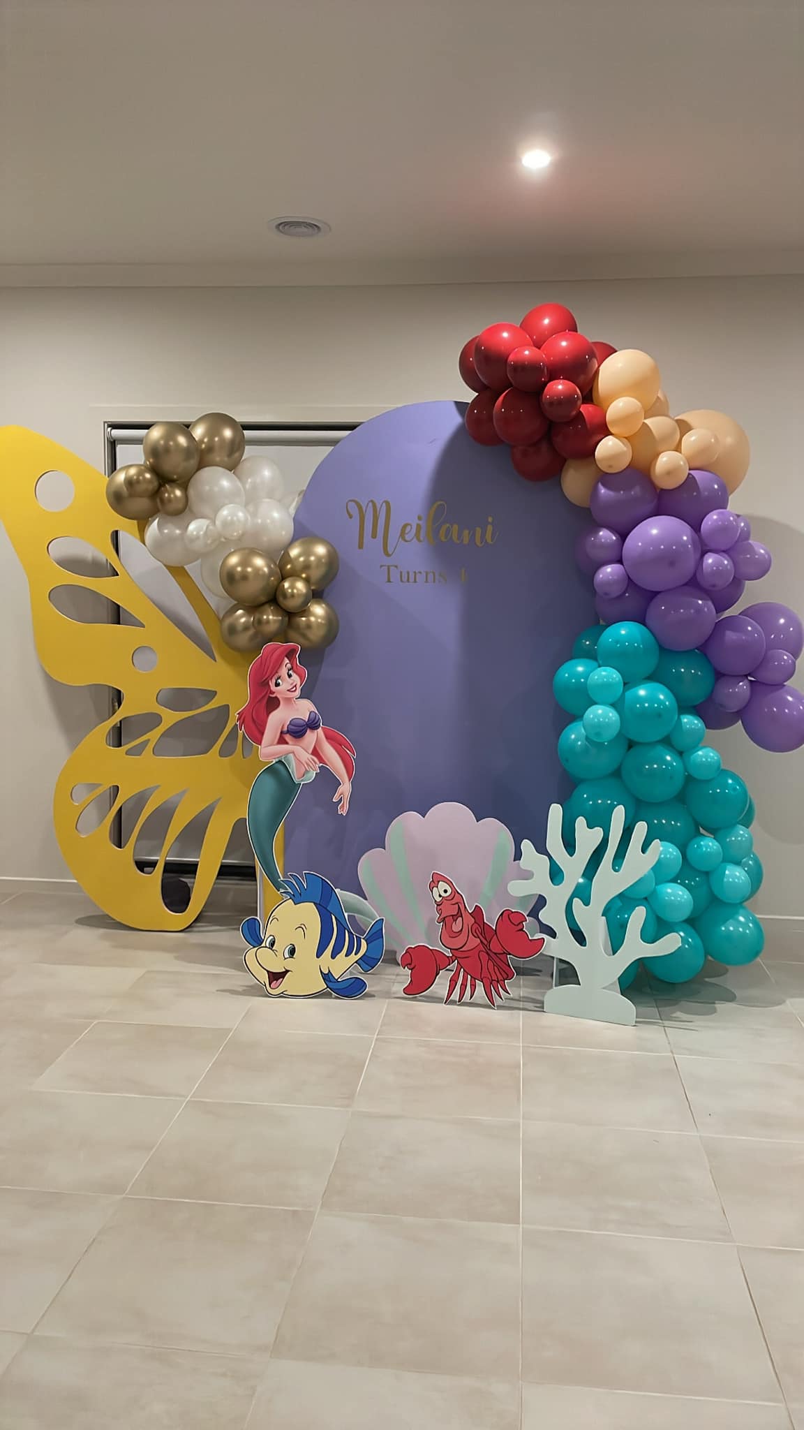Little Mermaid themed party backdrop featuring balloons, custom signage, and character cutouts of Ariel and Flounder.