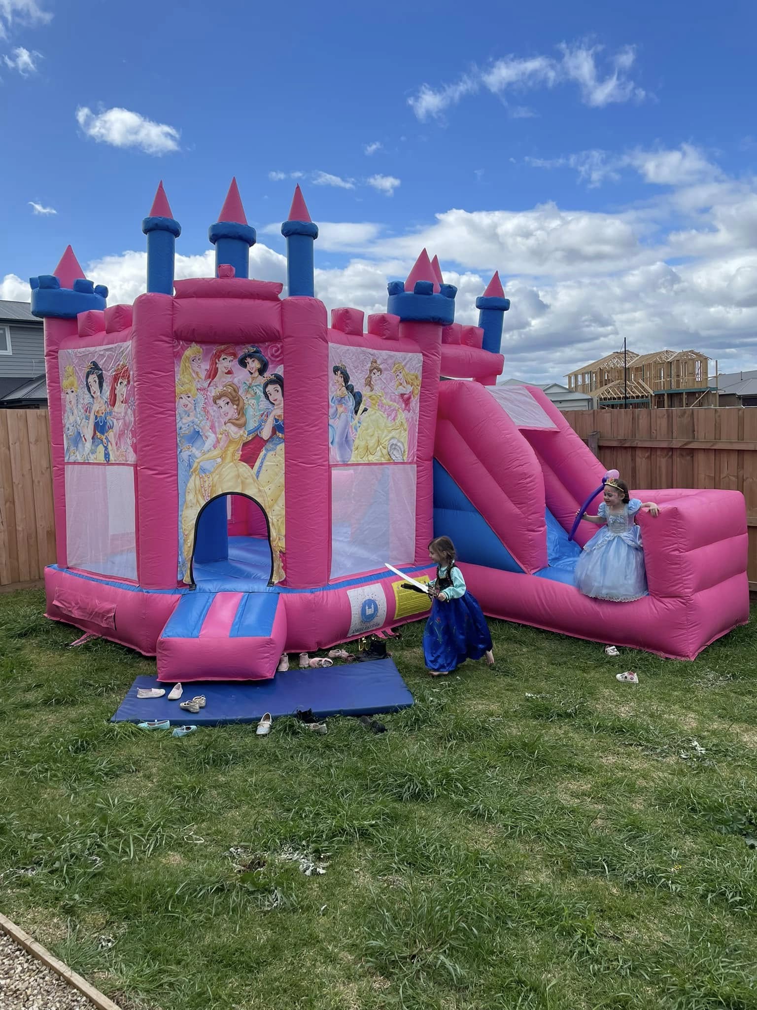 Pink and blue princess-themed inflatable bounce house with slide and two children in costumes playing outdoors
