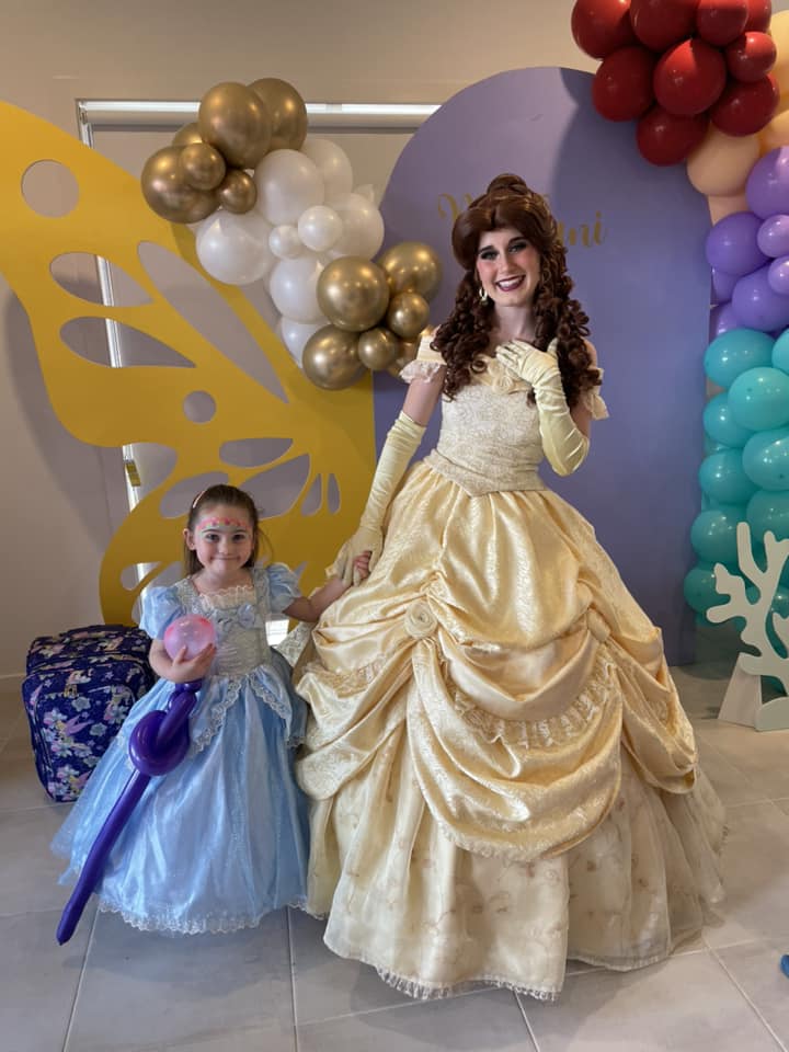 A young girl dressed as Cinderella holding a balloon wand standing next to a professional Belle princess performer.