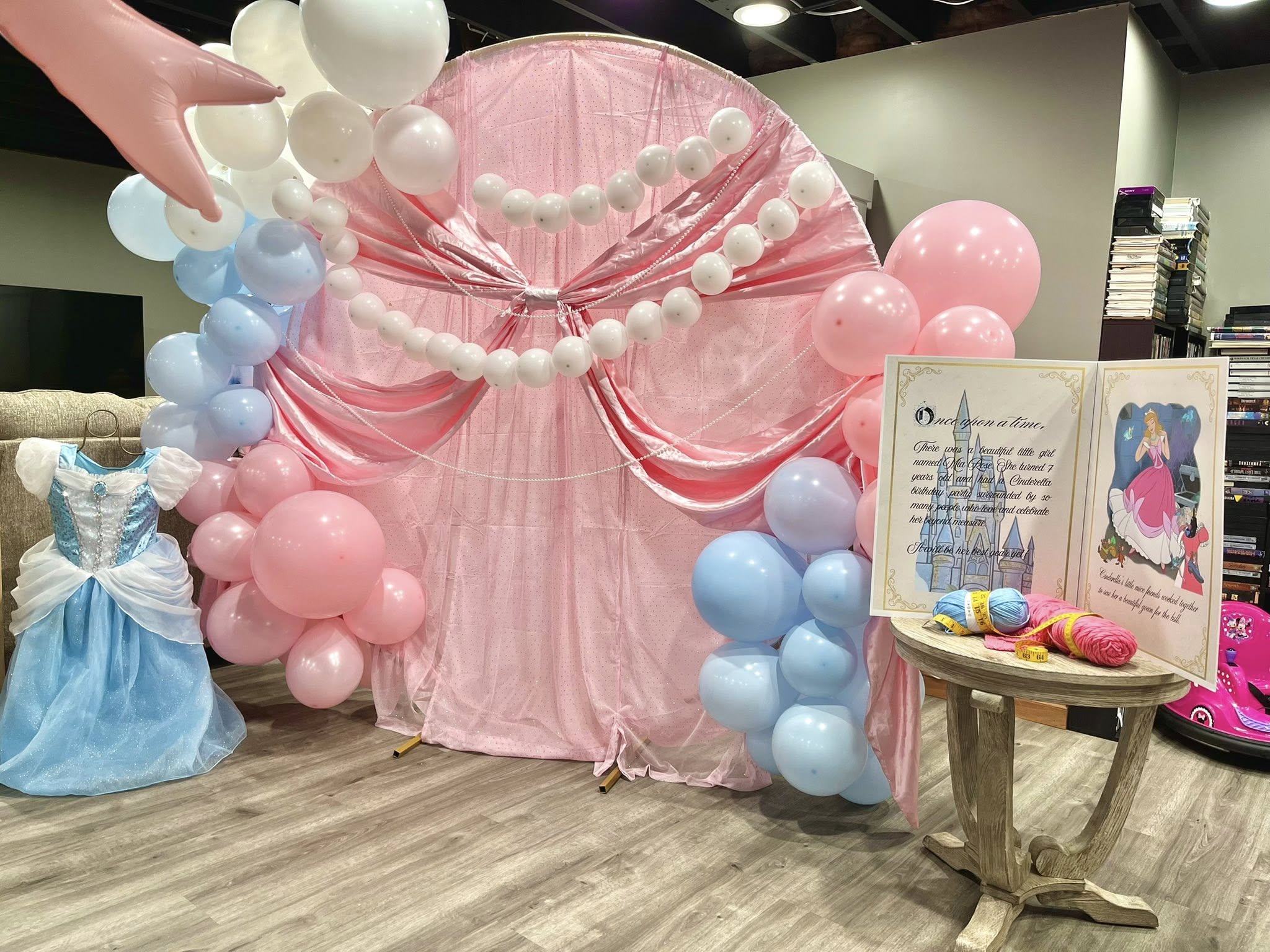 Cinderella themed party backdrop with pink fabric, balloon arches, a childs blue ballgown, and a storybook display