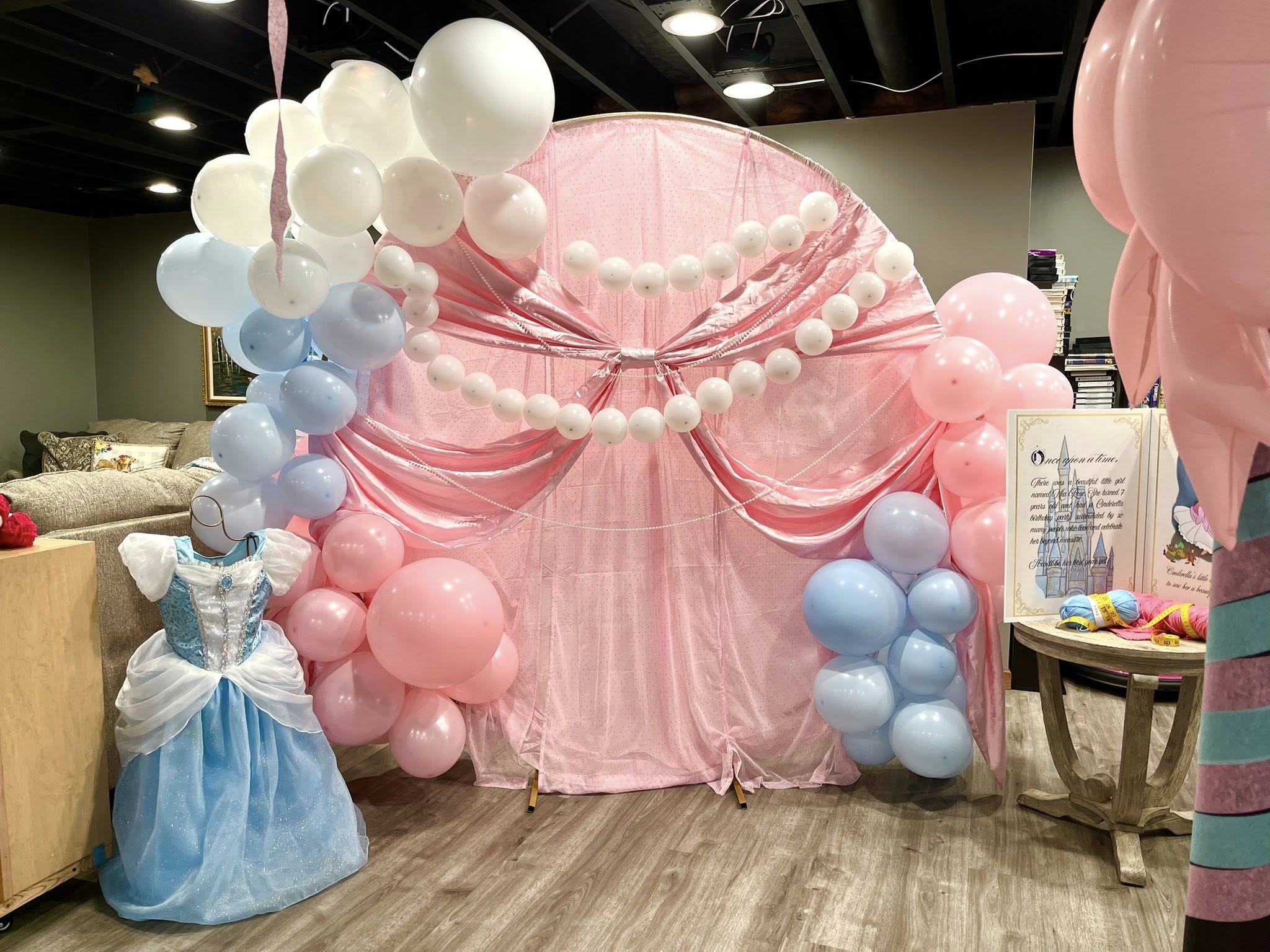 Cinderella-themed birthday party backdrop with pink fabric, white and blue balloons, and a princess dress display.