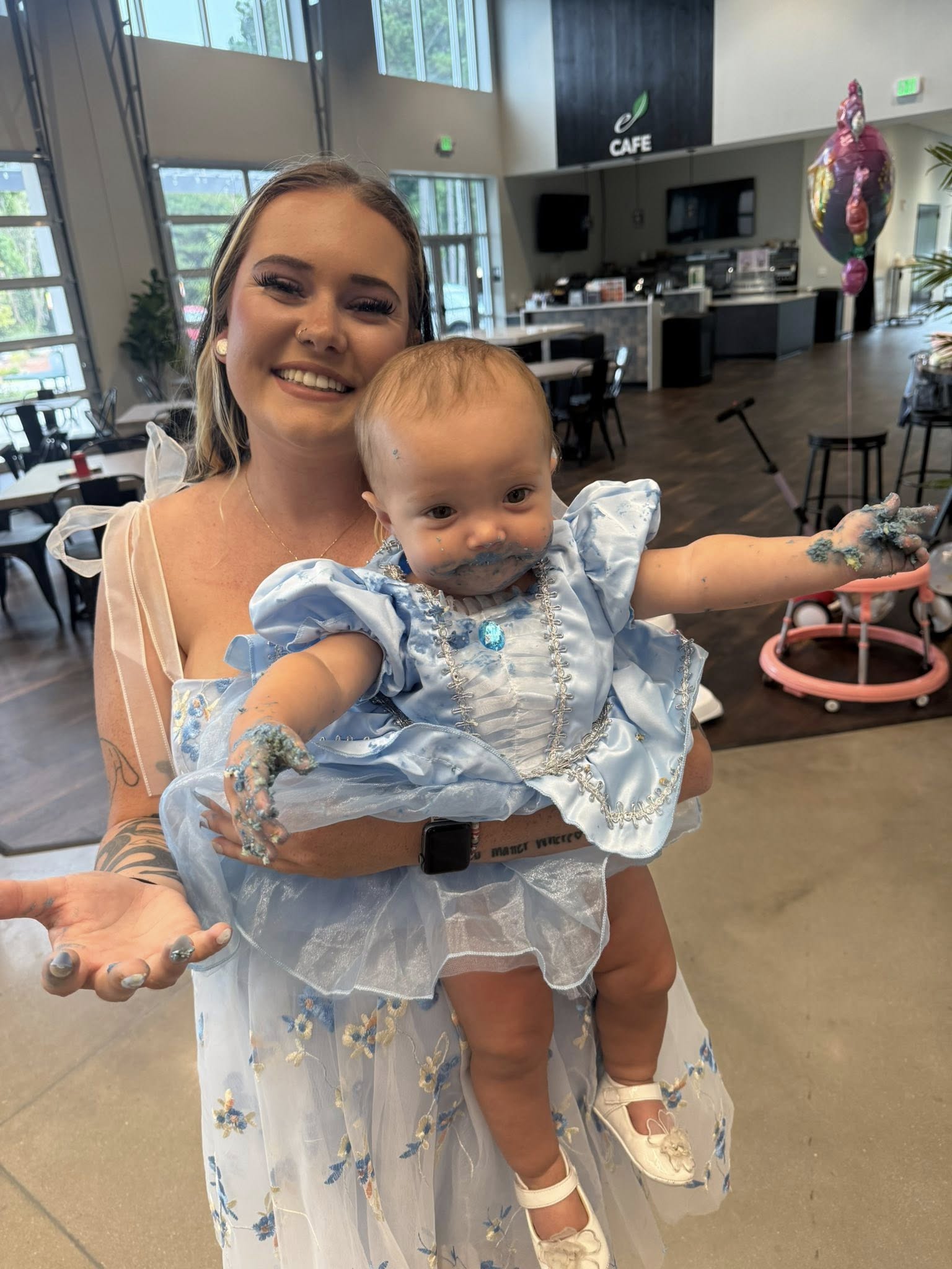 A smiling young woman holding her baby who is wearing a blue princess dress covered in frosting.