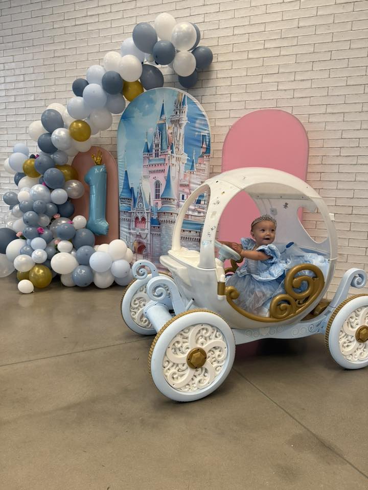 Baby in a Cinderella princess dress sitting in a carriage prop at a first birthday party with castle backdrop.