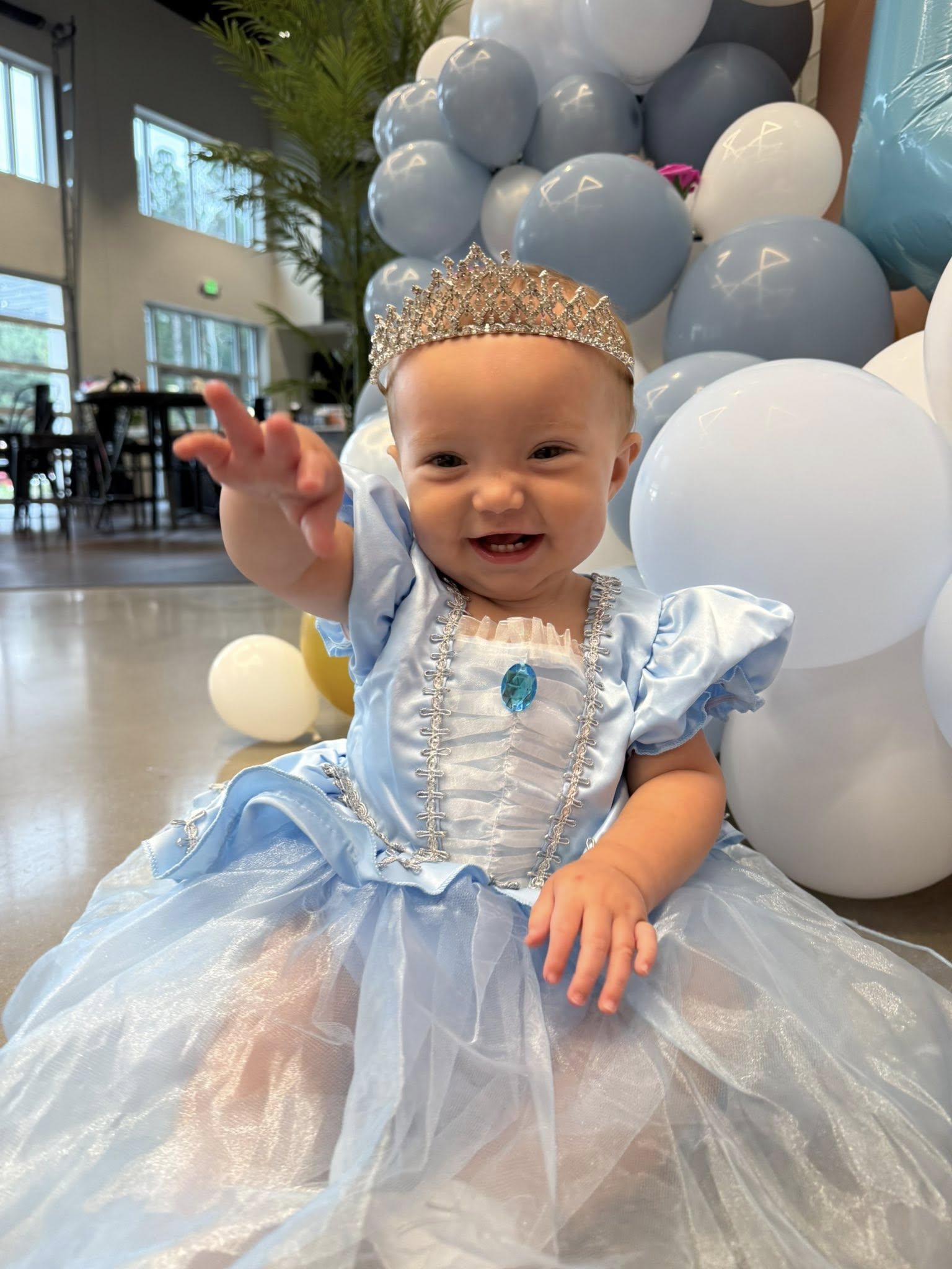 Baby smiling and reaching forward while wearing a blue princess costume and tiara amidst blue and white balloons