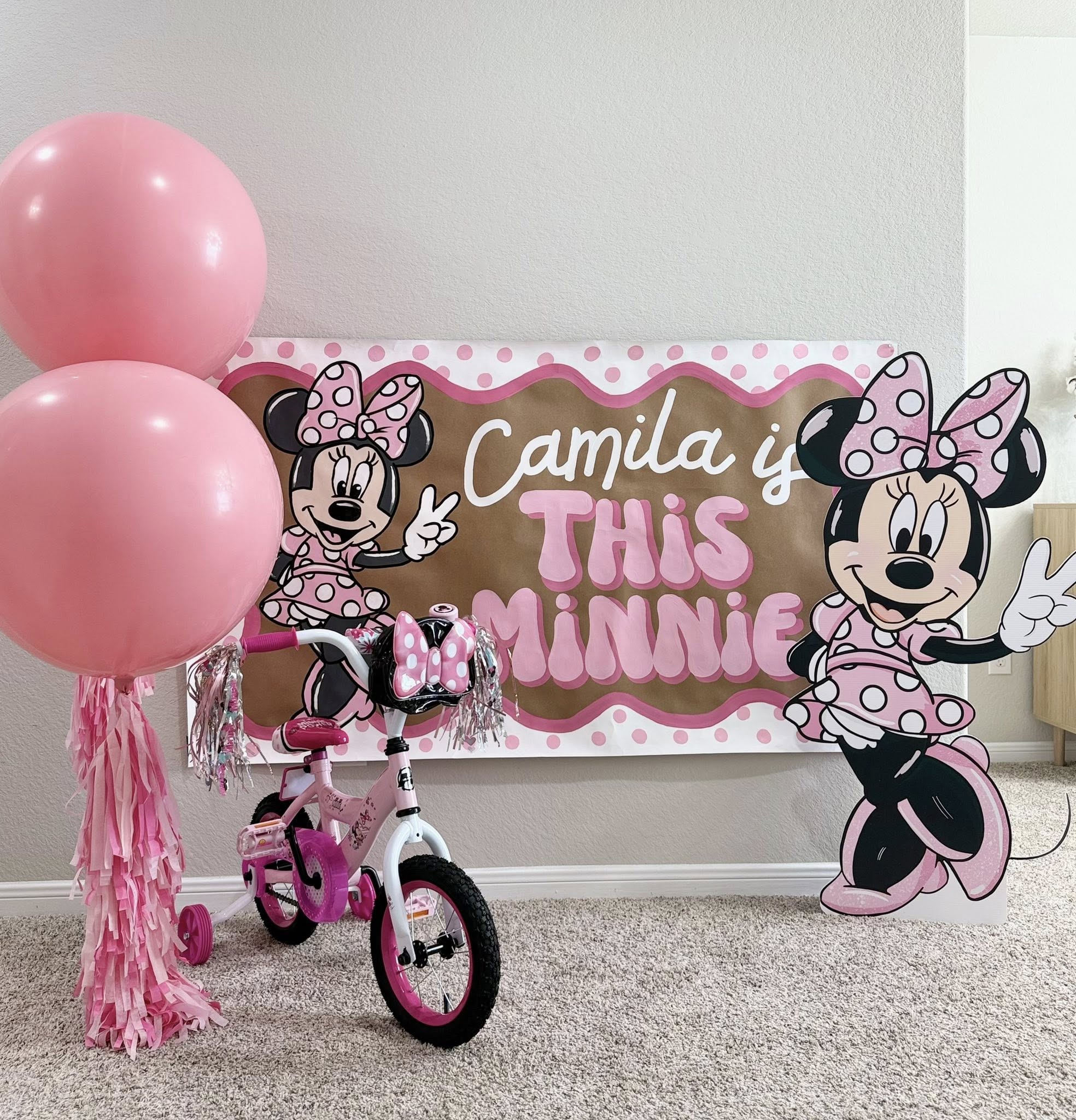 Minnie Mouse themed birthday party banner reading Camila is This Minnie with a pink bicycle and balloons.