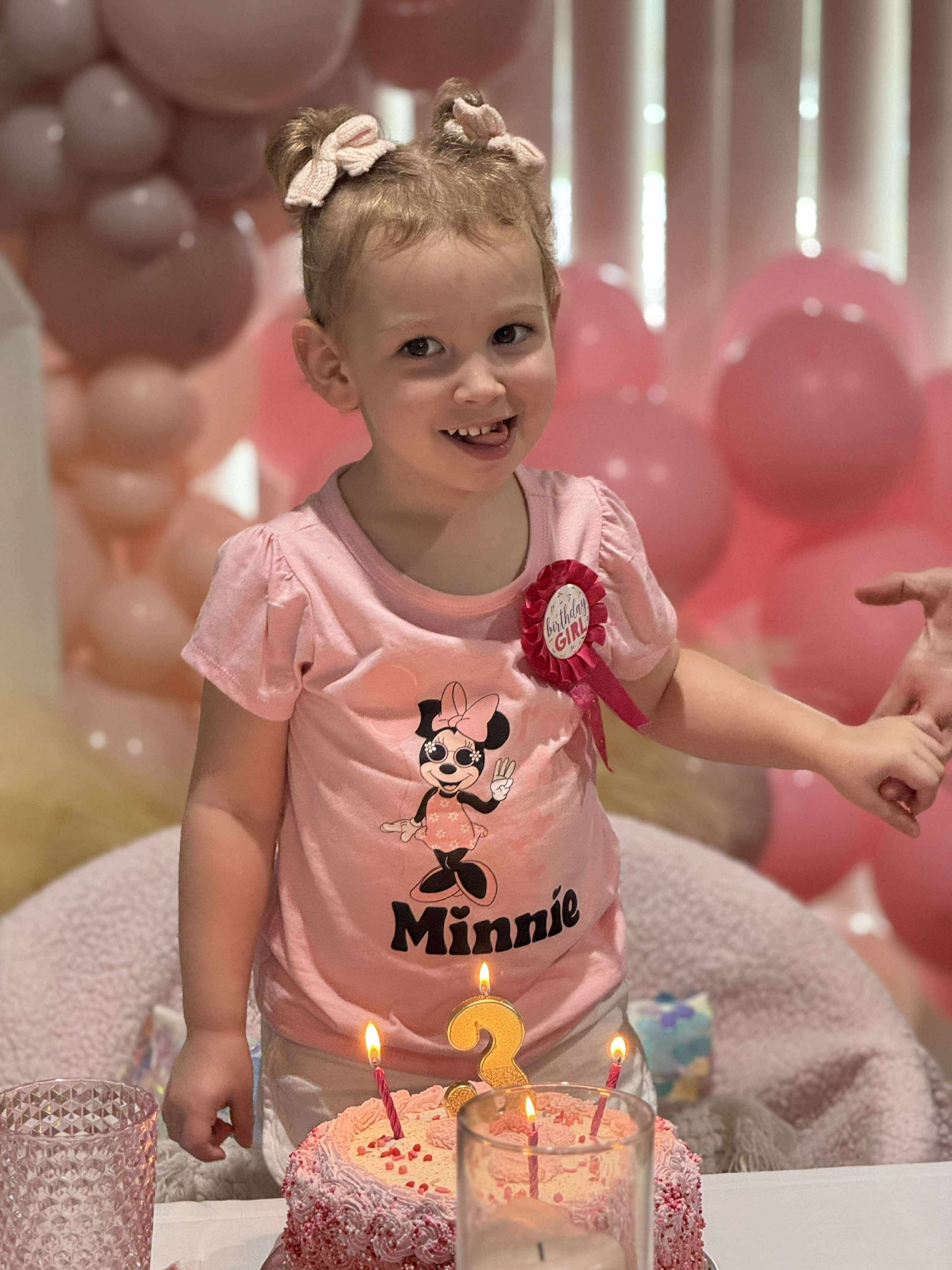 Toddler wearing a pink Minnie Mouse shirt and Birthday Girl badge in front of a second birthday cake.