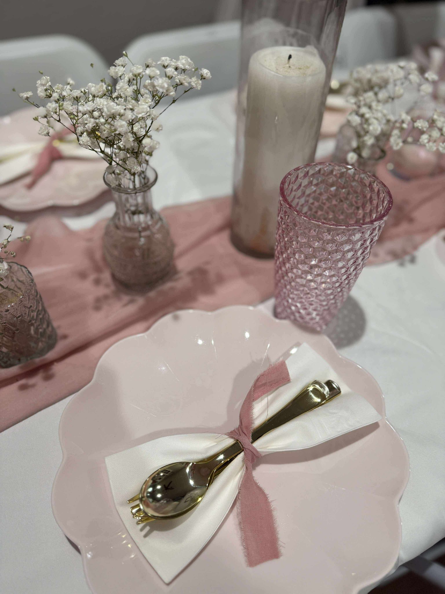 Table setting featuring a scalloped pink plate, white napkin with pink ribbon, gold cutlery, and floral centerpiece.