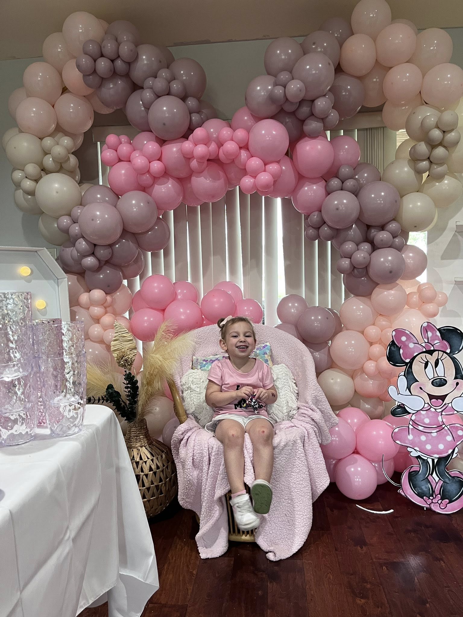 A smiling young child sitting in a chair decorated for a birthday party with a pink balloon arch and Minnie Mouse cutout.