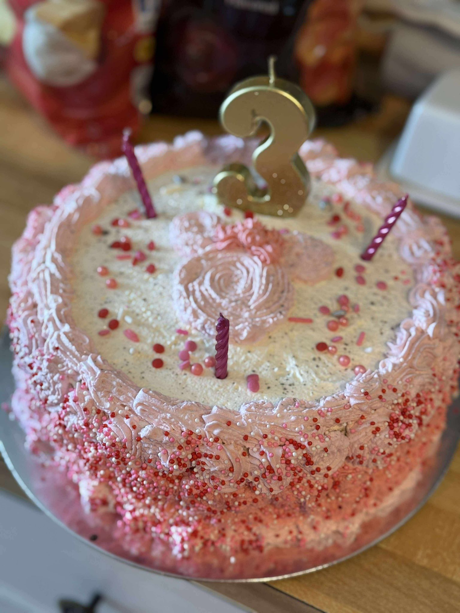 A pink birthday cake decorated with sprinkles and a golden number 3 candle.