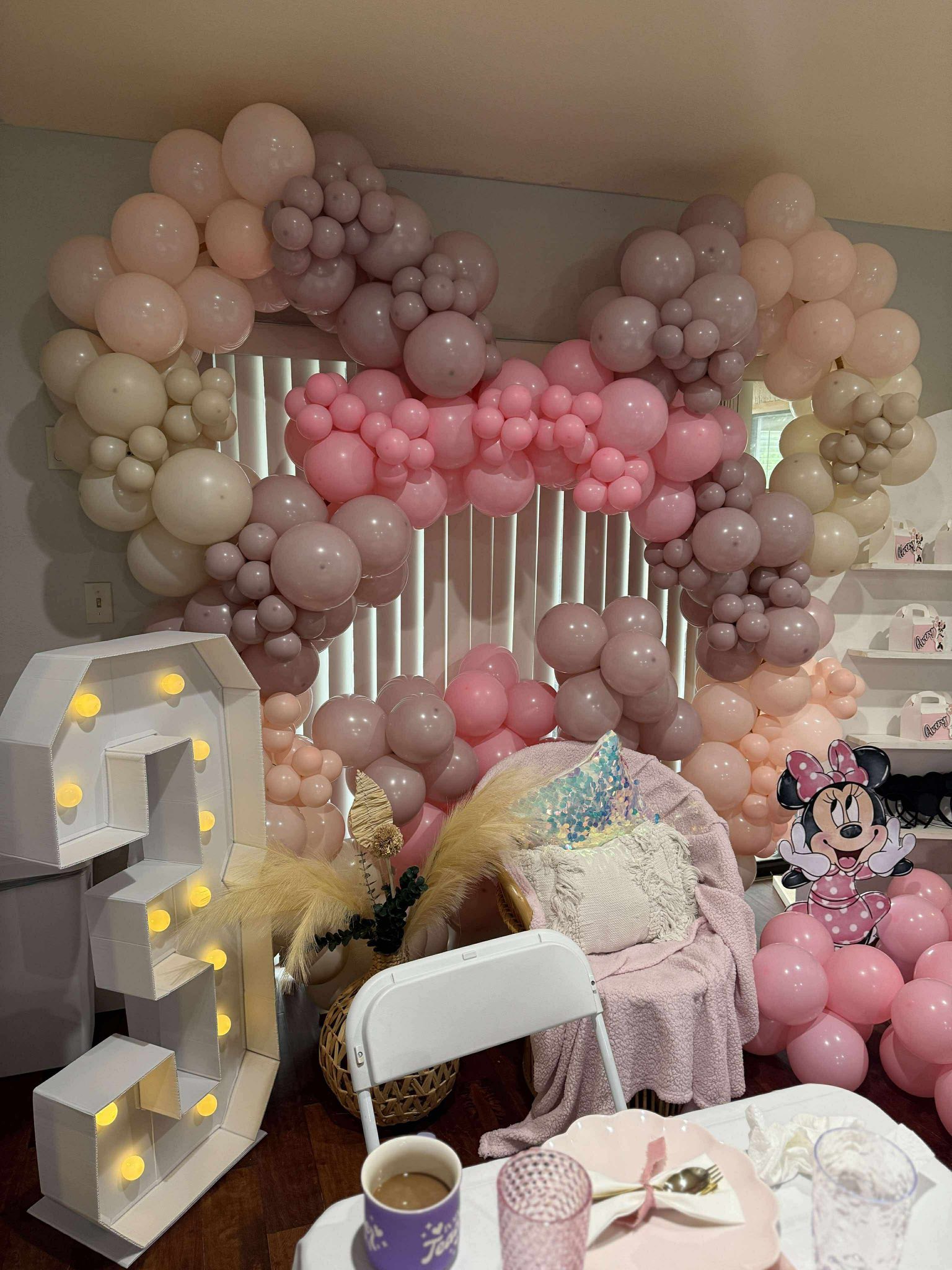Boho birthday party decor with a white light-up number 3, pink and mauve balloon arch, and Minnie Mouse cutout.