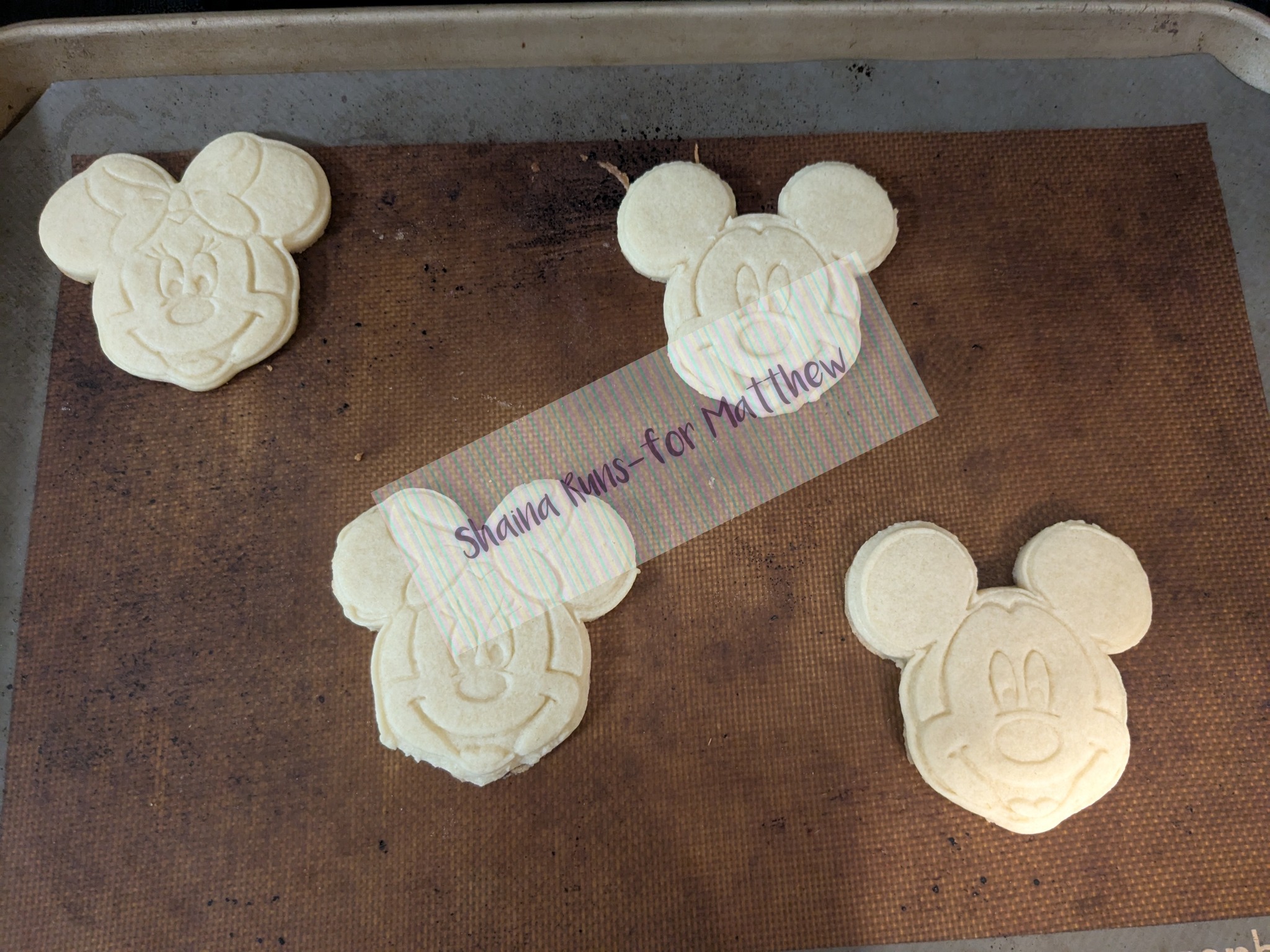 Four baked Mickey Mouse shaped sugar cookies on a baking sheet.